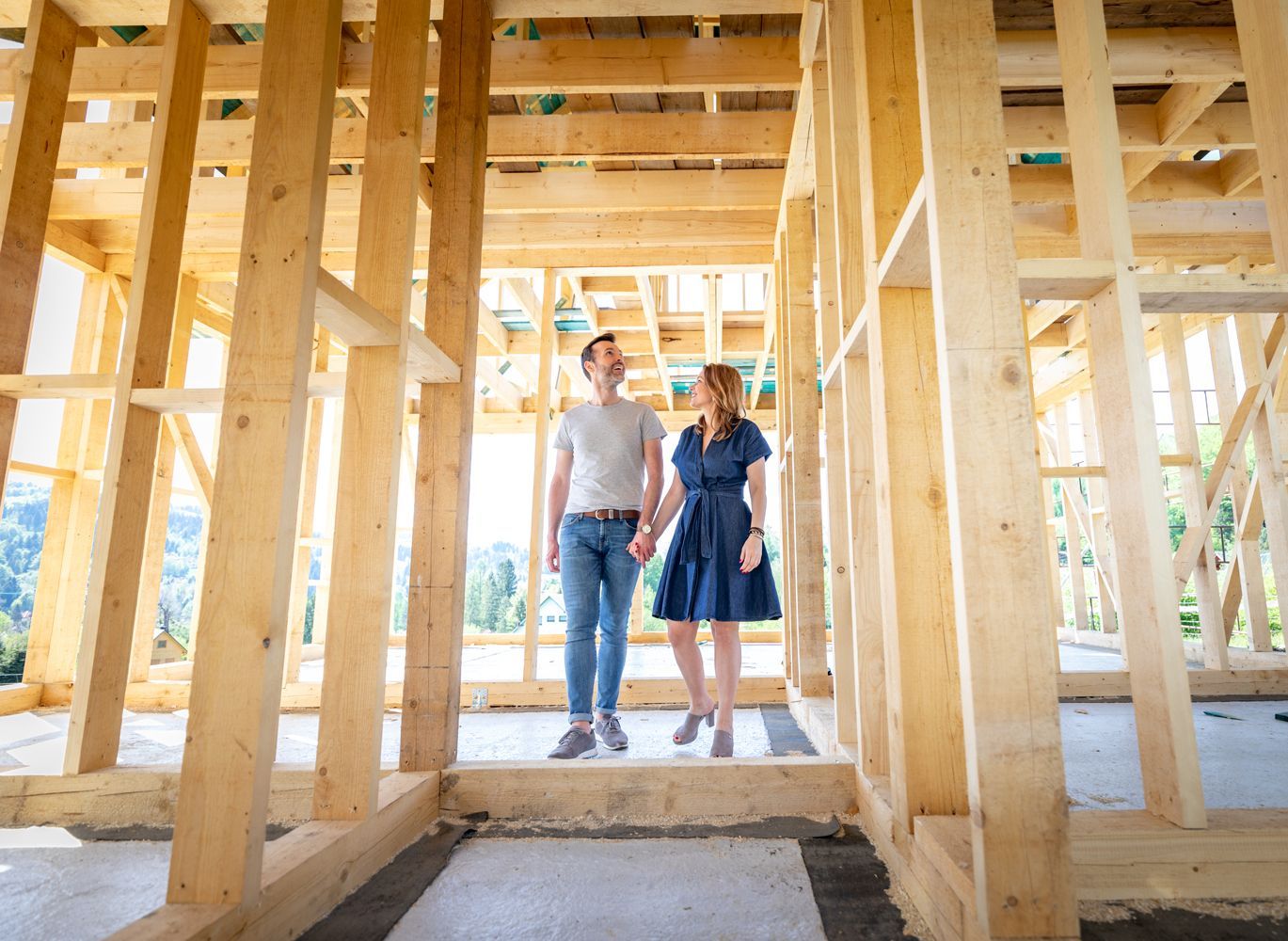 A couple walking through the wooden framework of a house under construction, illuminated by bright daylight.