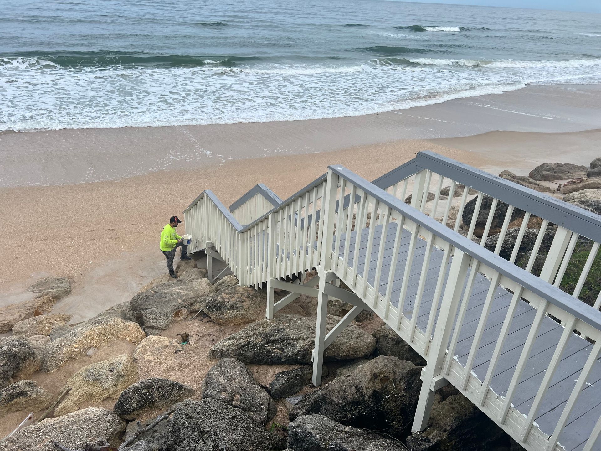 A worker in a high-visibility jacket stands on a sandy beach next to a wooden staircase descending over a rocky shoreline.