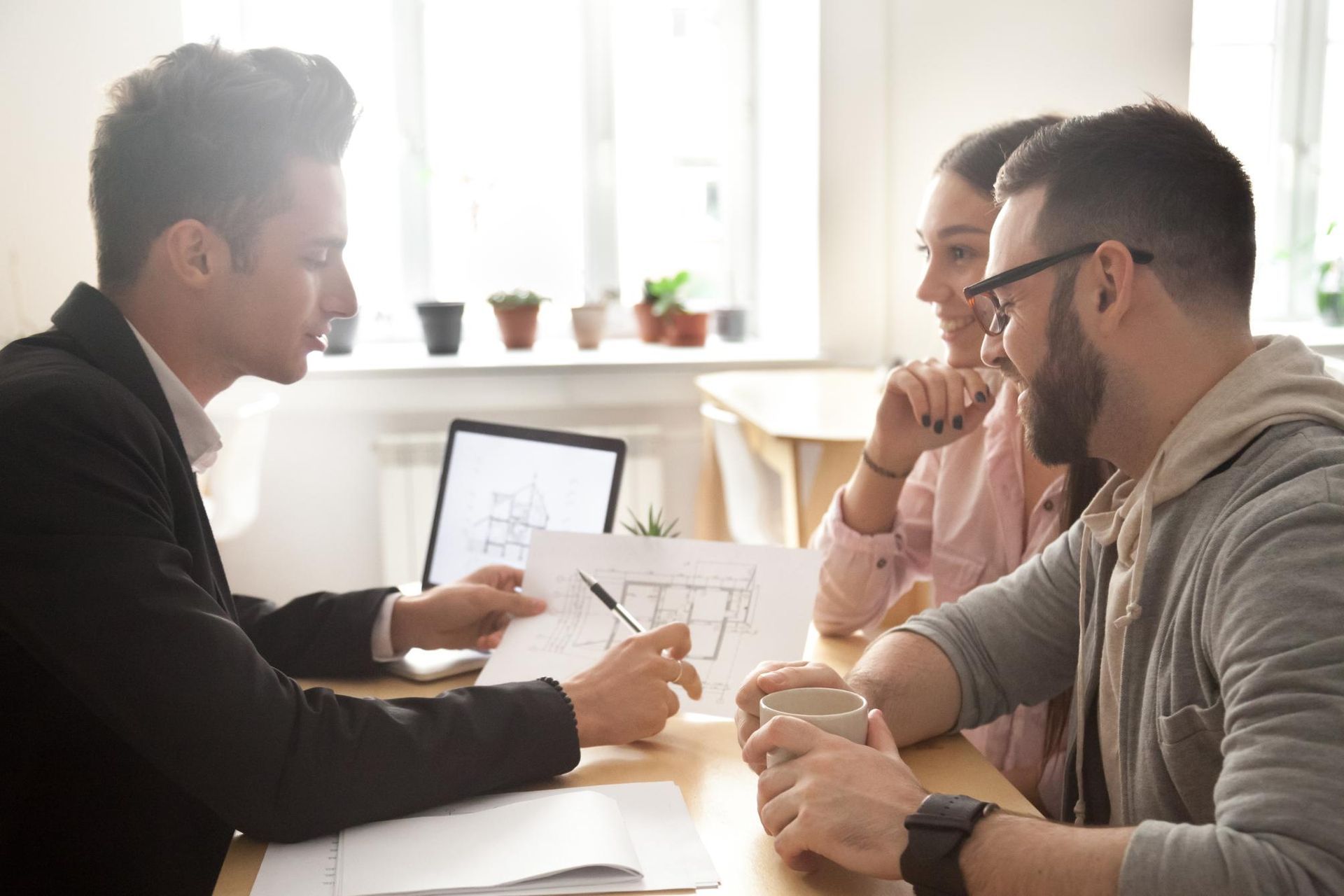 A professional explains a floor plan to a couple sitting across from them at a wooden desk in a bright office.