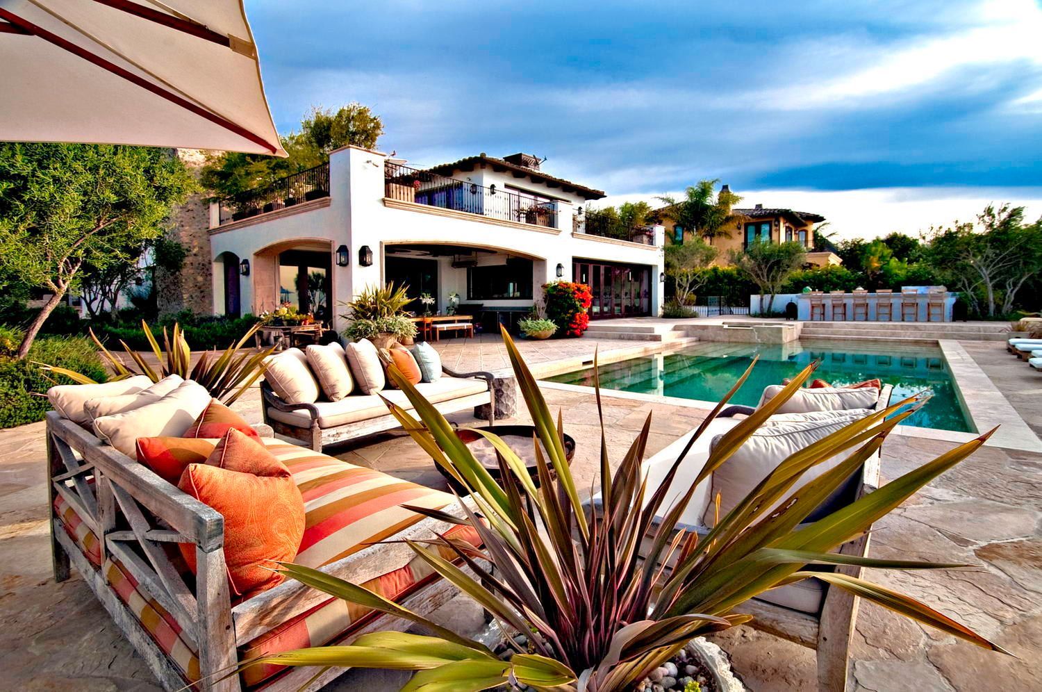 A luxurious villa patio with outdoor lounge seating, a swimming pool, and tropical plants under a cloudy blue sky.
