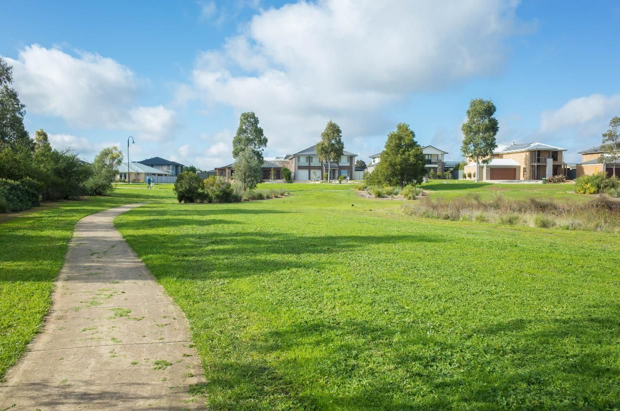 A paved path curves through a grassy field toward houses in a residential neighborhood under a bright, sunny blue sky.