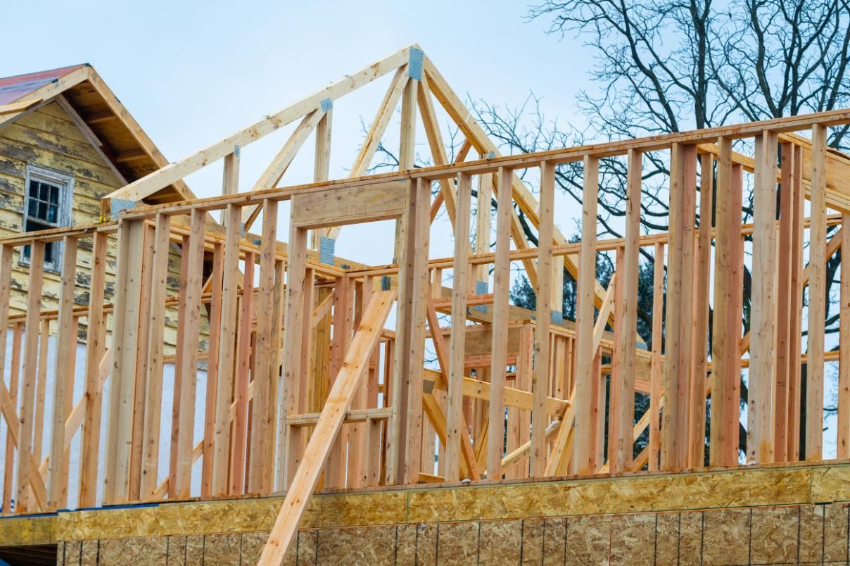 The wood frame of a house under construction, featuring exposed wall studs and roof trusses against a light sky.