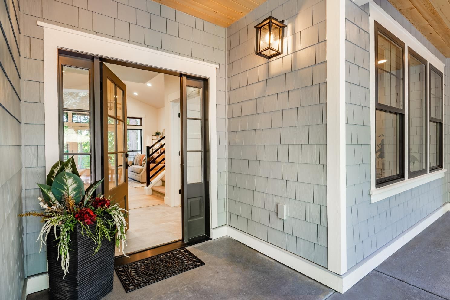 An open front door leads into a home, framed by grey shake siding, a hanging lantern, and a potted plant on the porch.