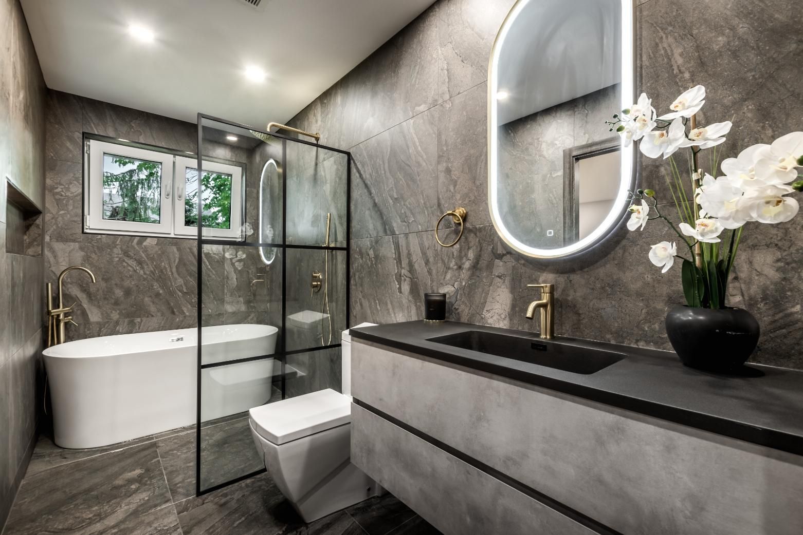 Modern bathroom with grey stone walls, a white freestanding tub, a walk-in glass shower, and a minimalist vanity.