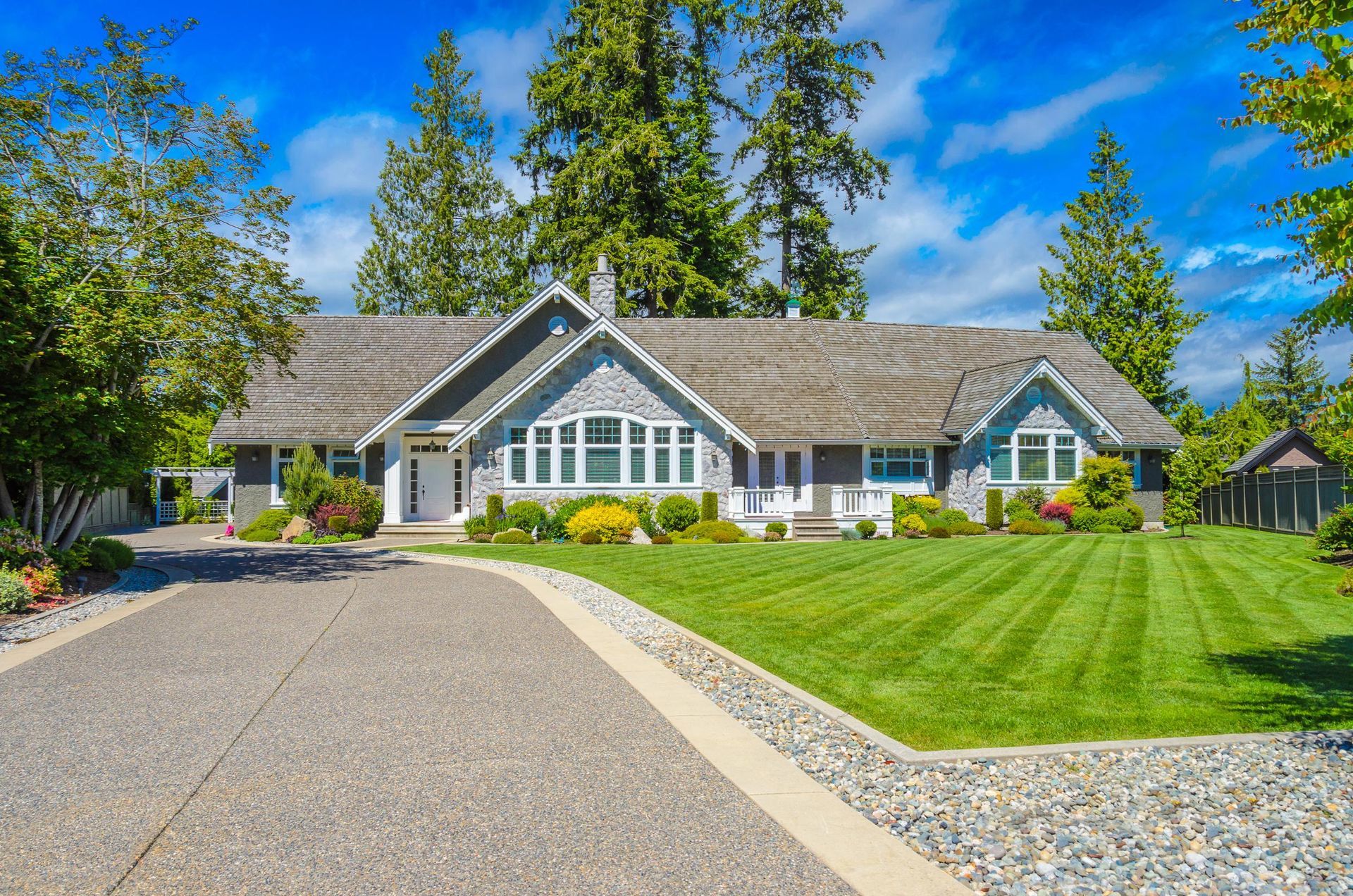 A gray house with a peaked roof and white trim sits behind a large, striped lawn and a long gravel driveway.