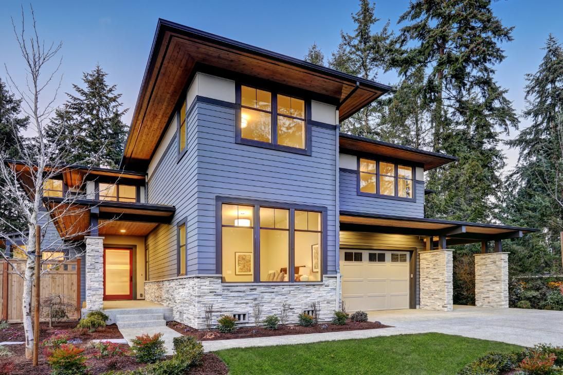 A two-story blue-sided modern house with a stone base, wooden eaves, and an attached garage surrounded by pine trees.