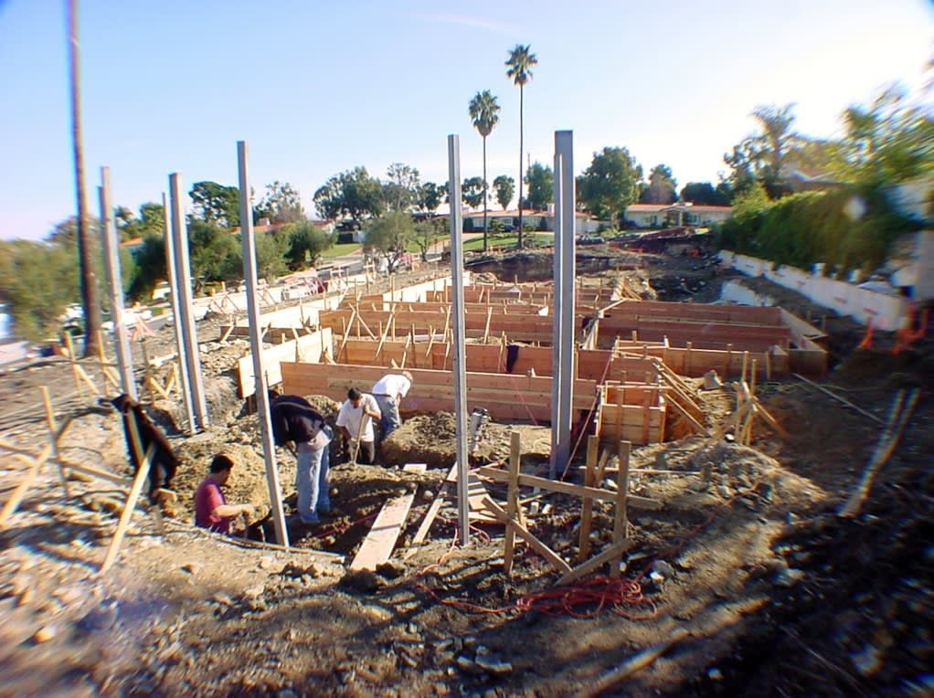 Construction workers build wooden forms for a concrete foundation, surrounded by tall steel columns in a dirt lot.