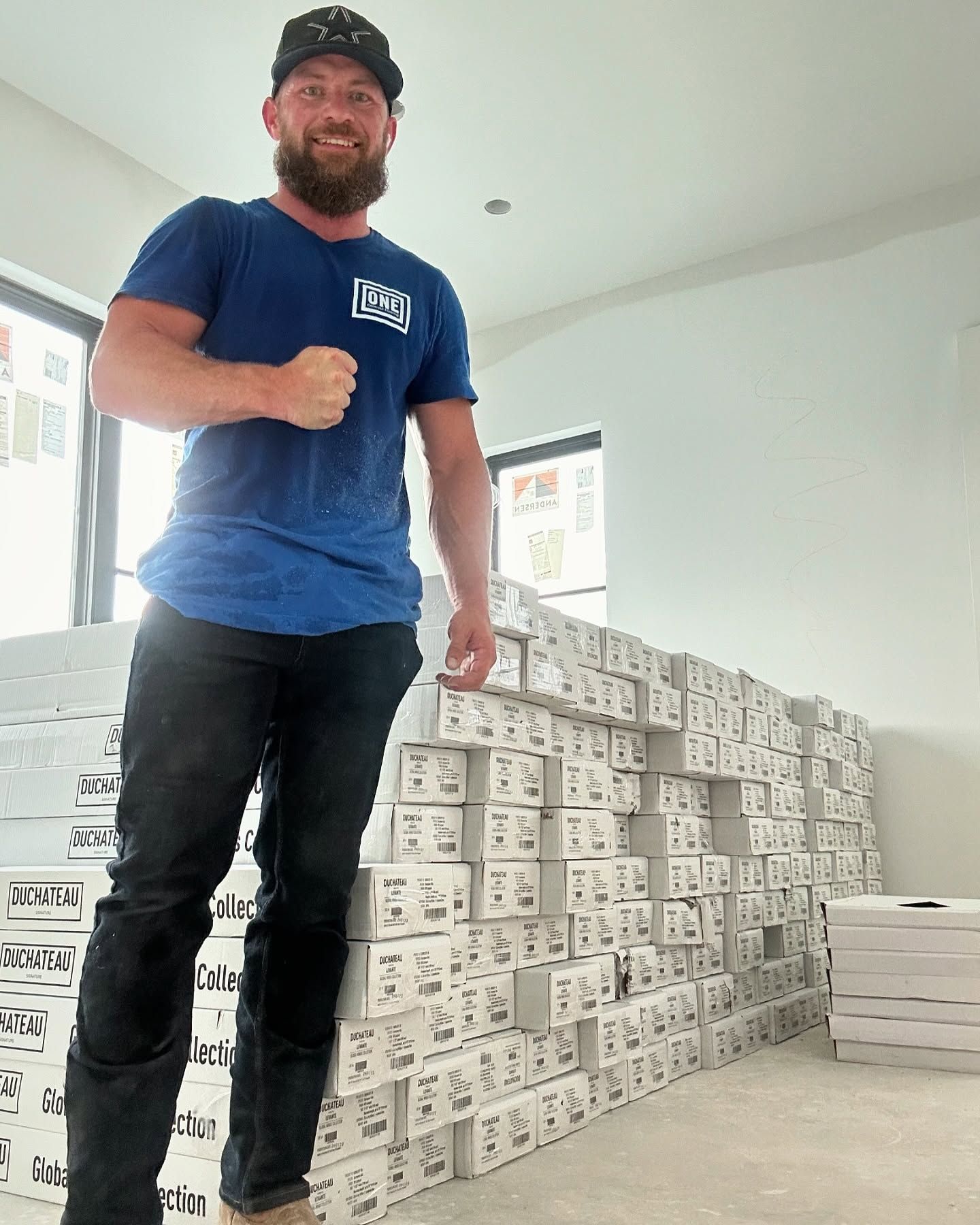 Man in blue shirt and black pants with clenched fist next to a large stack of boxes in a room.