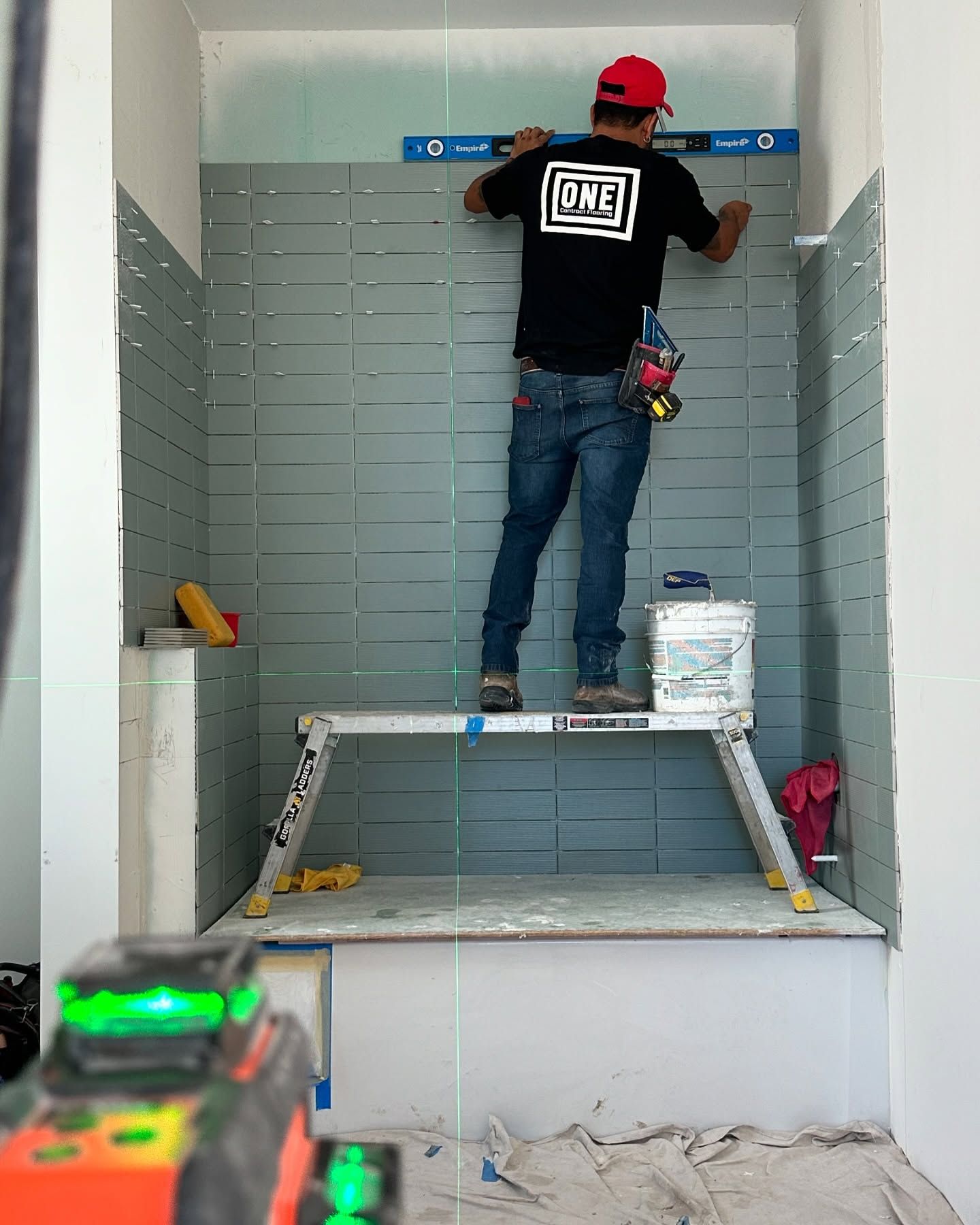 Man using level, tiling bathroom wall. He stands on a platform. Construction tools are visible.