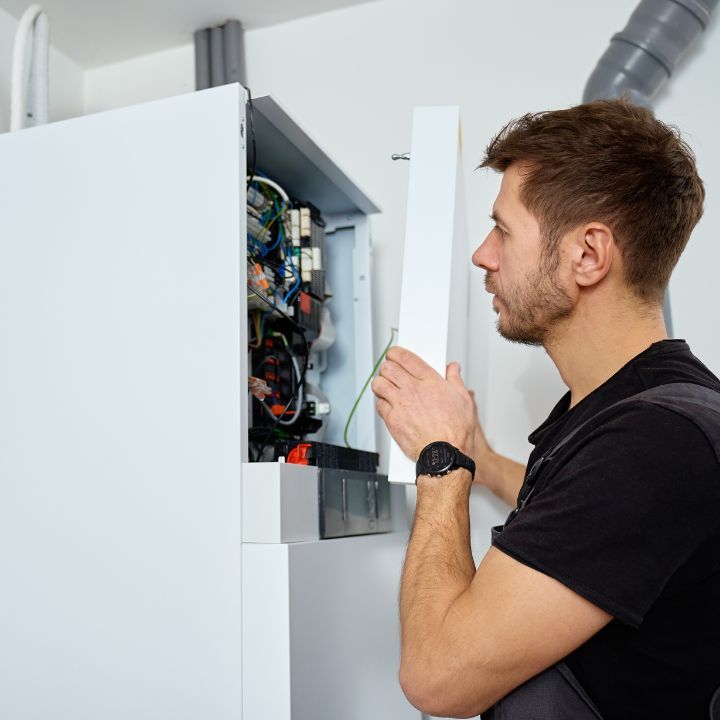 A person wearing a black shirt opens the white metal panel of a wall-mounted boiler to inspect the internal components.
