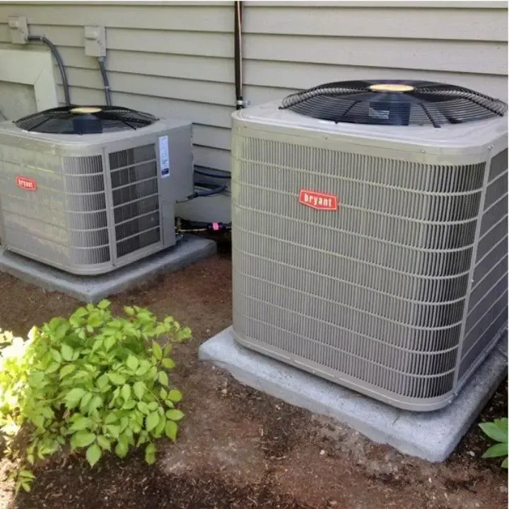 Two Bryant HVAC outdoor condenser units sit on concrete pads beside a house with beige siding and a small green bush.