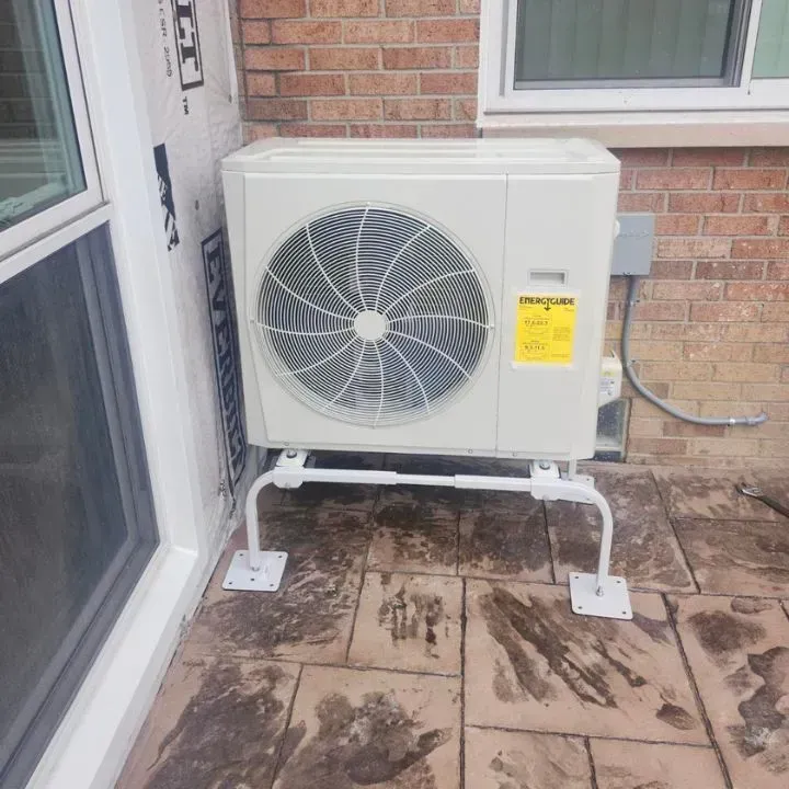 Outdoor air conditioning unit on a raised white stand beside a brick wall and patio door.
