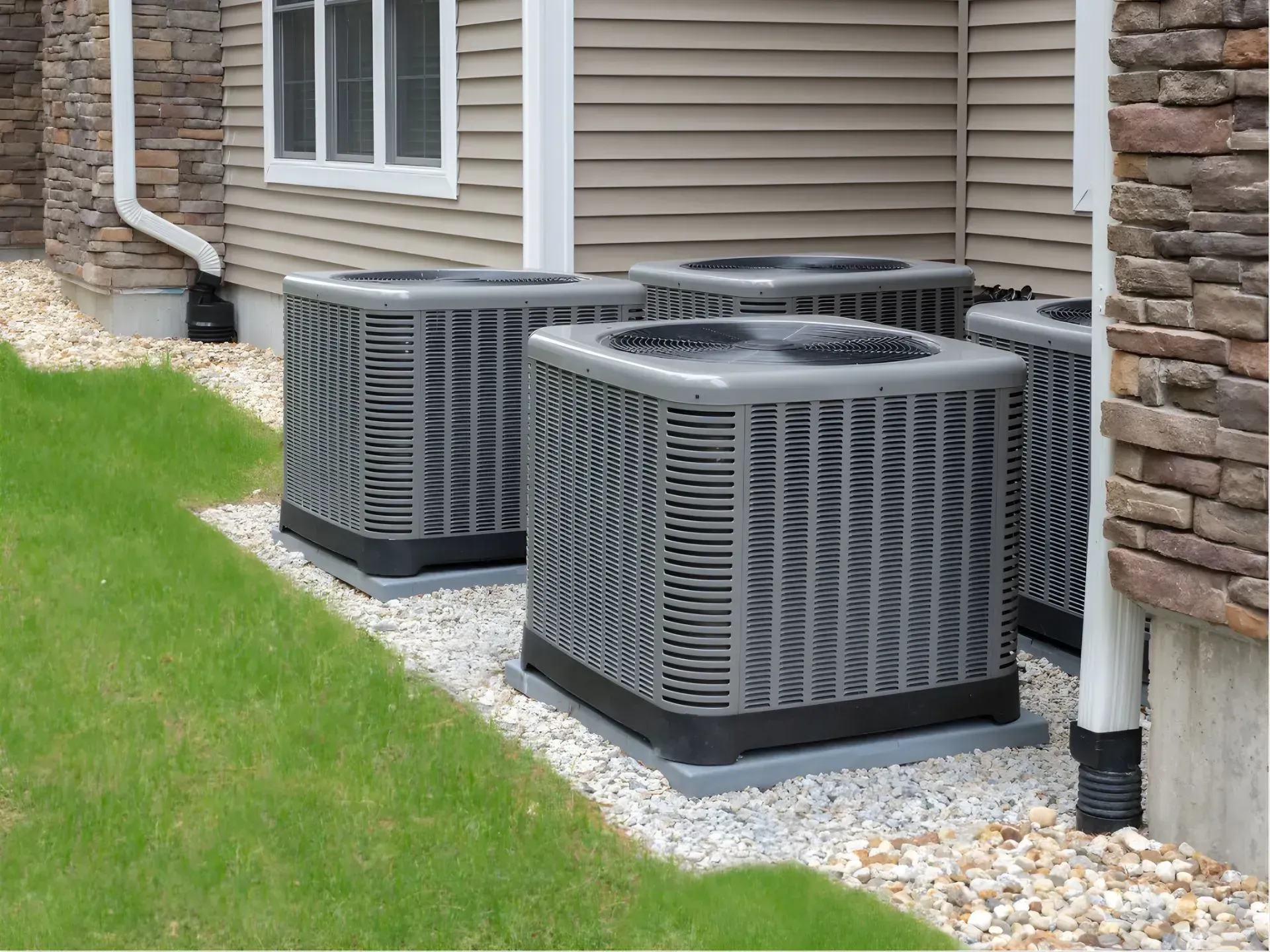 Two outdoor air conditioning units beside a house, next to a stone wall and green lawn.