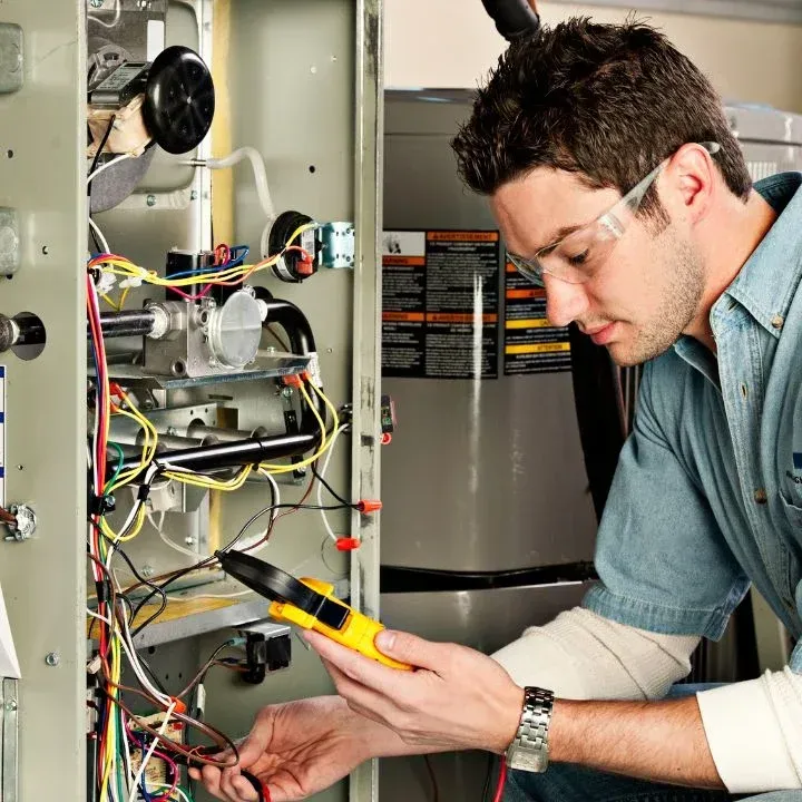 A technician in safety glasses uses a multimeter to inspect the wiring of an open furnace unit.
