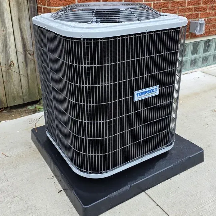 Outdoor air conditioner unit on a concrete pad beside a wooden fence and glass-block wall