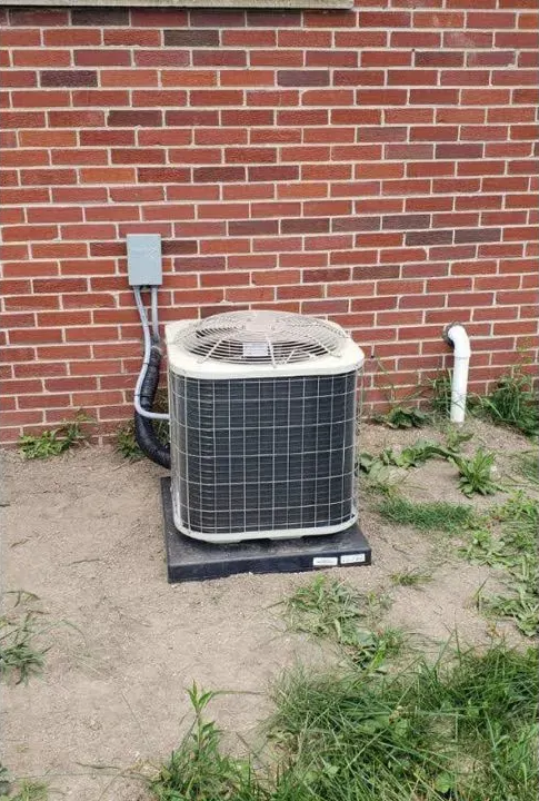 Outdoor air conditioner unit beside a red brick wall on a patchy dirt and grass yard