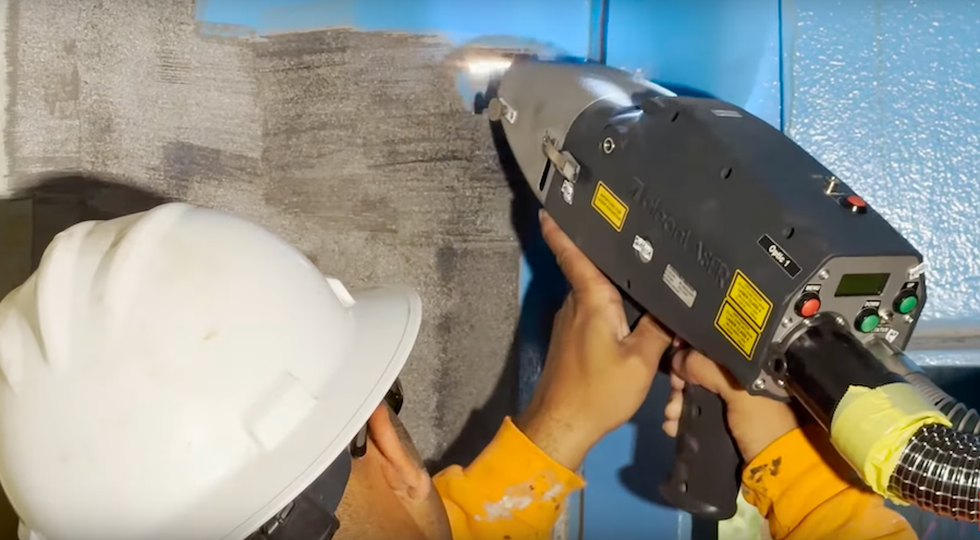 Person in hardhat using a laser device to remove coating from a wall.