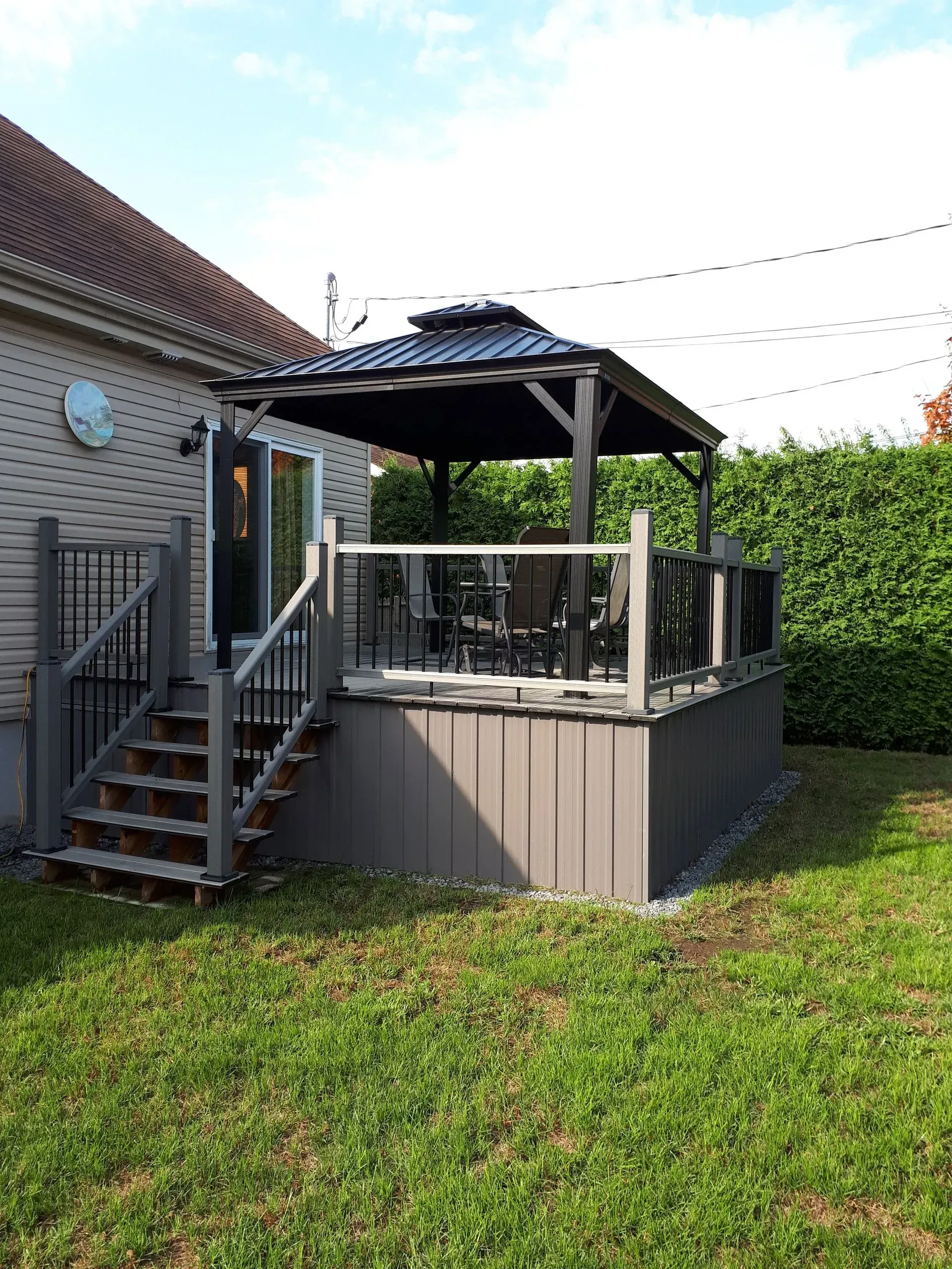 Terrasse arrière avec escalier, rambarde noire et gazebo couvert, jouxtant une maison et une haie.