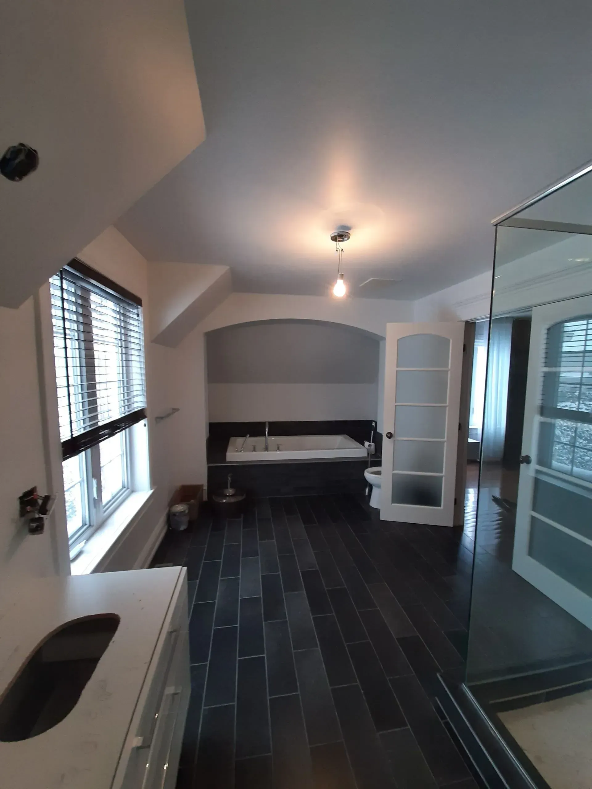 Salle de bains moderne avec carrelage foncé au sol, grande fenêtre, miroir mural et cabine de douche en verre.
