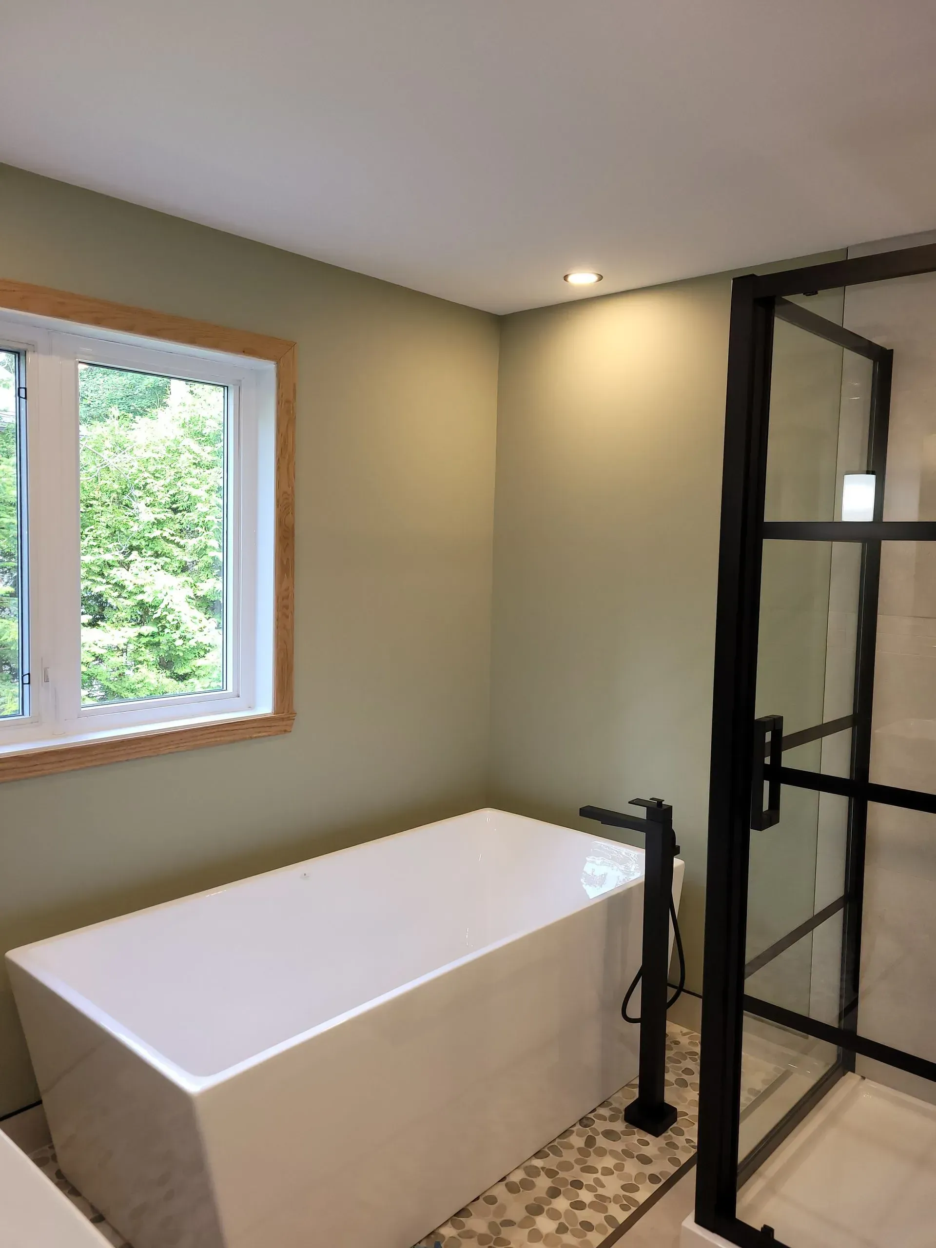 Salle de bains avec baignoire blanche, douche en verre à cadre noir, murs beiges et fenêtre à encadrement en bois.