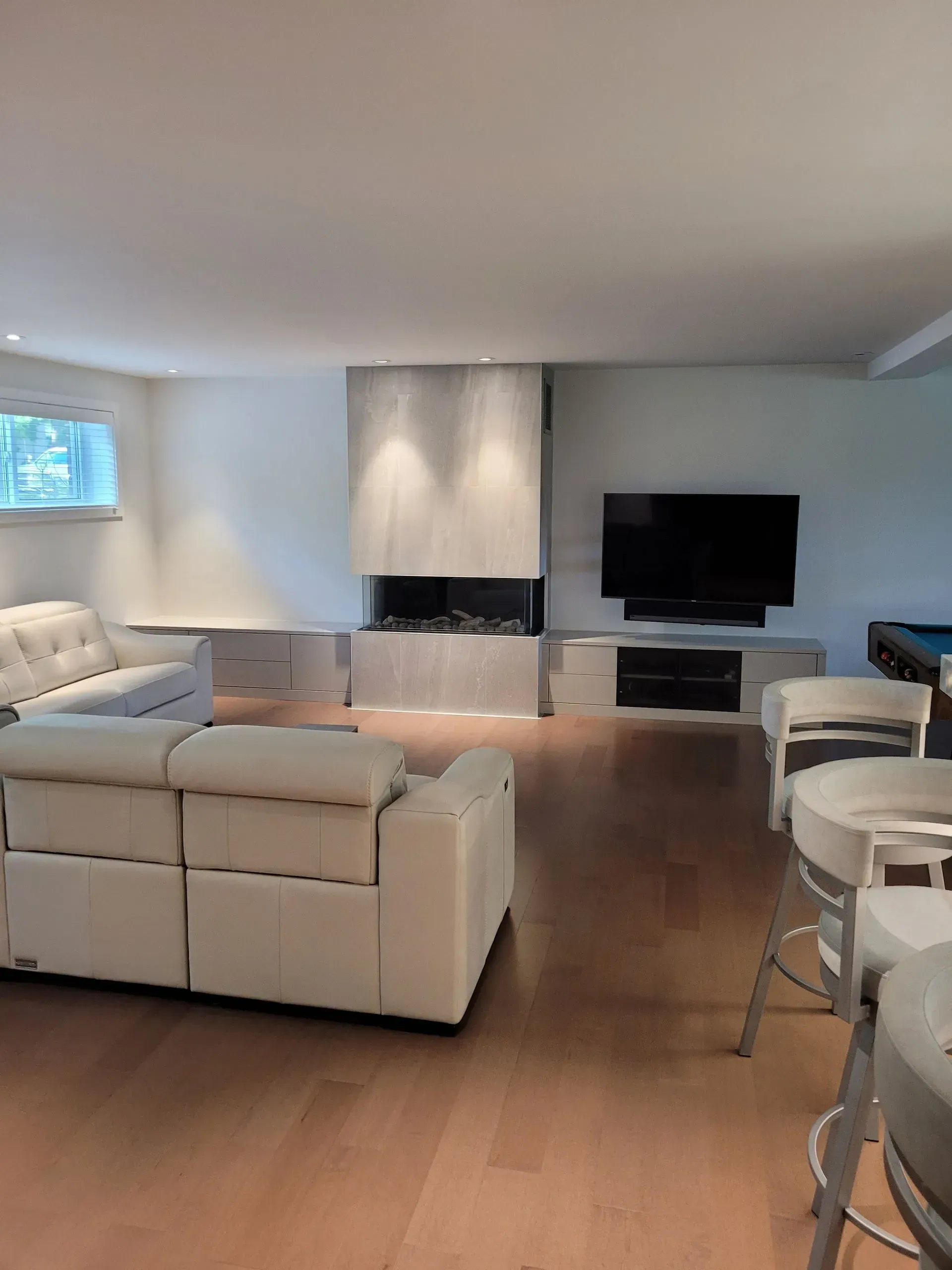 Salon moderne avec canapés blancs, parquet, télévision murale et grandes fenêtres.
