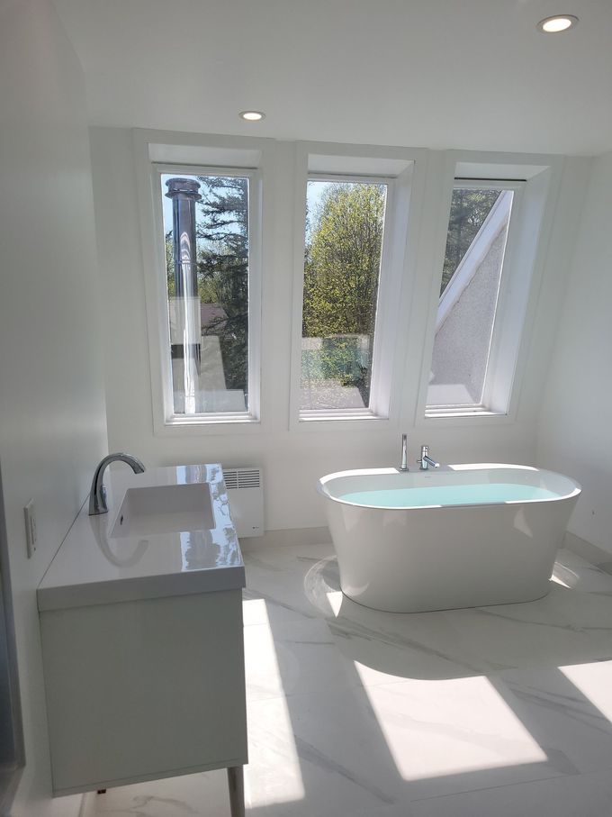 Salle de bains moderne et lumineuse avec baignoire îlot, meuble-lavabo et hautes fenêtres laissant entrer la lumière du soleil.