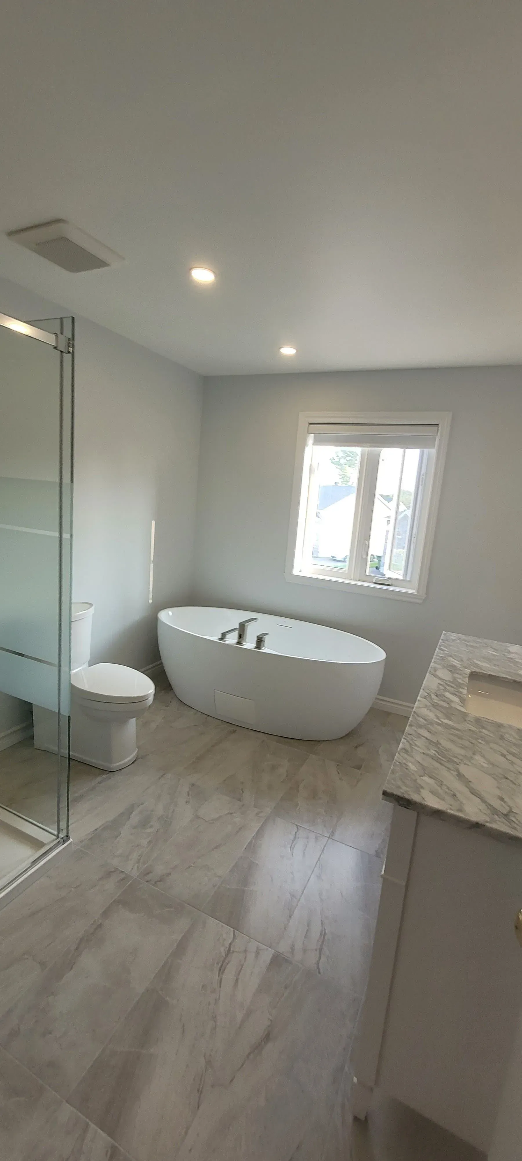 Salle de bains moderne et lumineuse avec baignoire îlot, douche en verre, meuble-lavabo et éclairage par fenêtre.