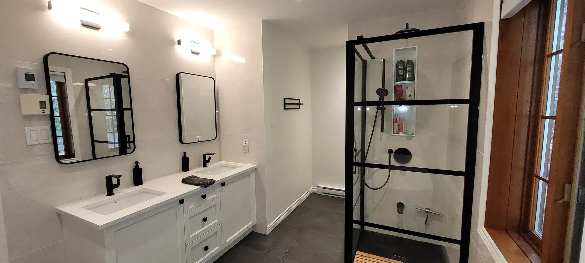 Salle de bains moderne avec double vasque, miroirs à cadre noir et cabine de douche en verre derrière une porte en bois.