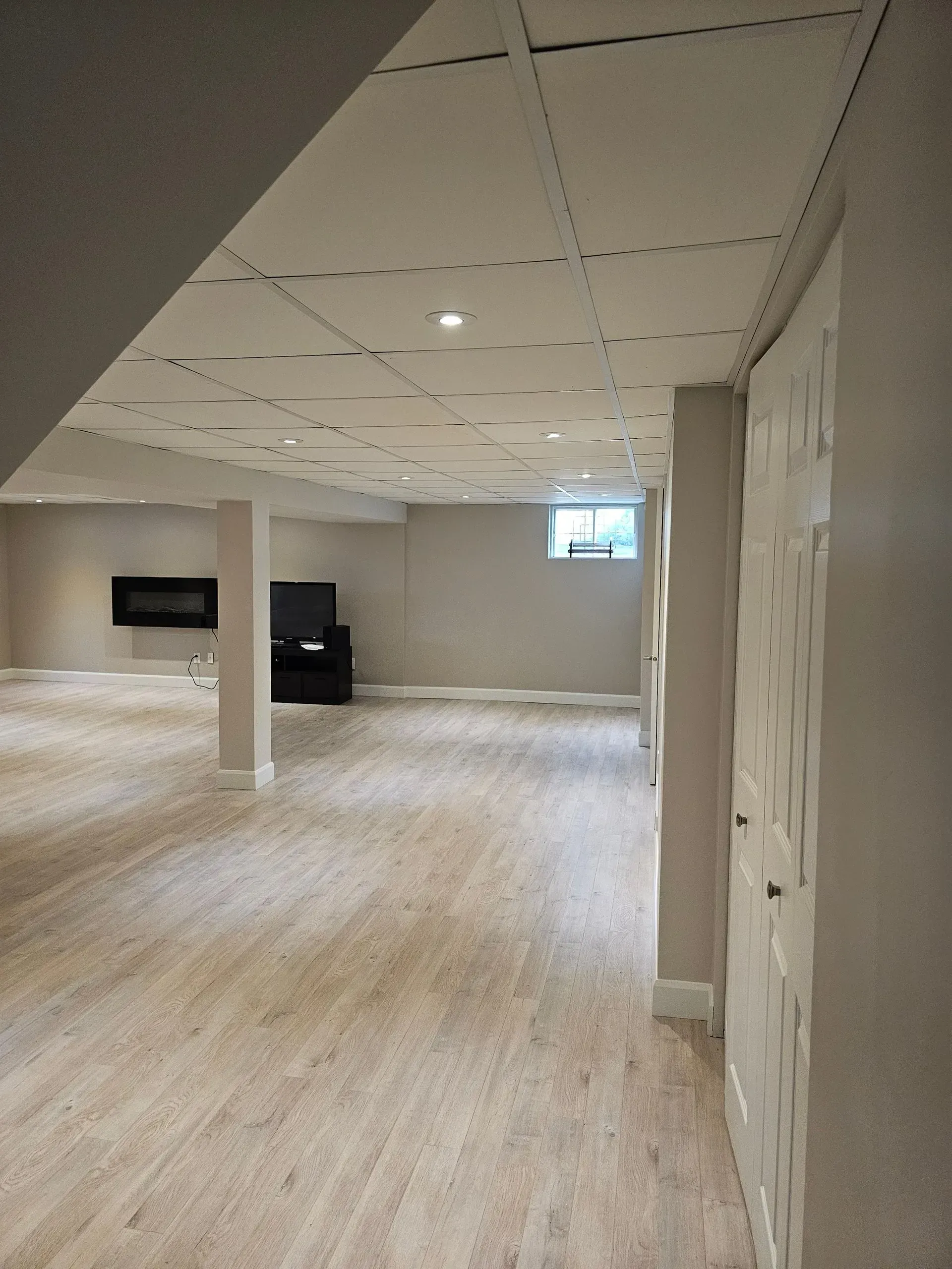 Pièce vide et inachevée avec des murs beiges, un sol imitation bois, un faux plafond et une télévision fixée au mur.