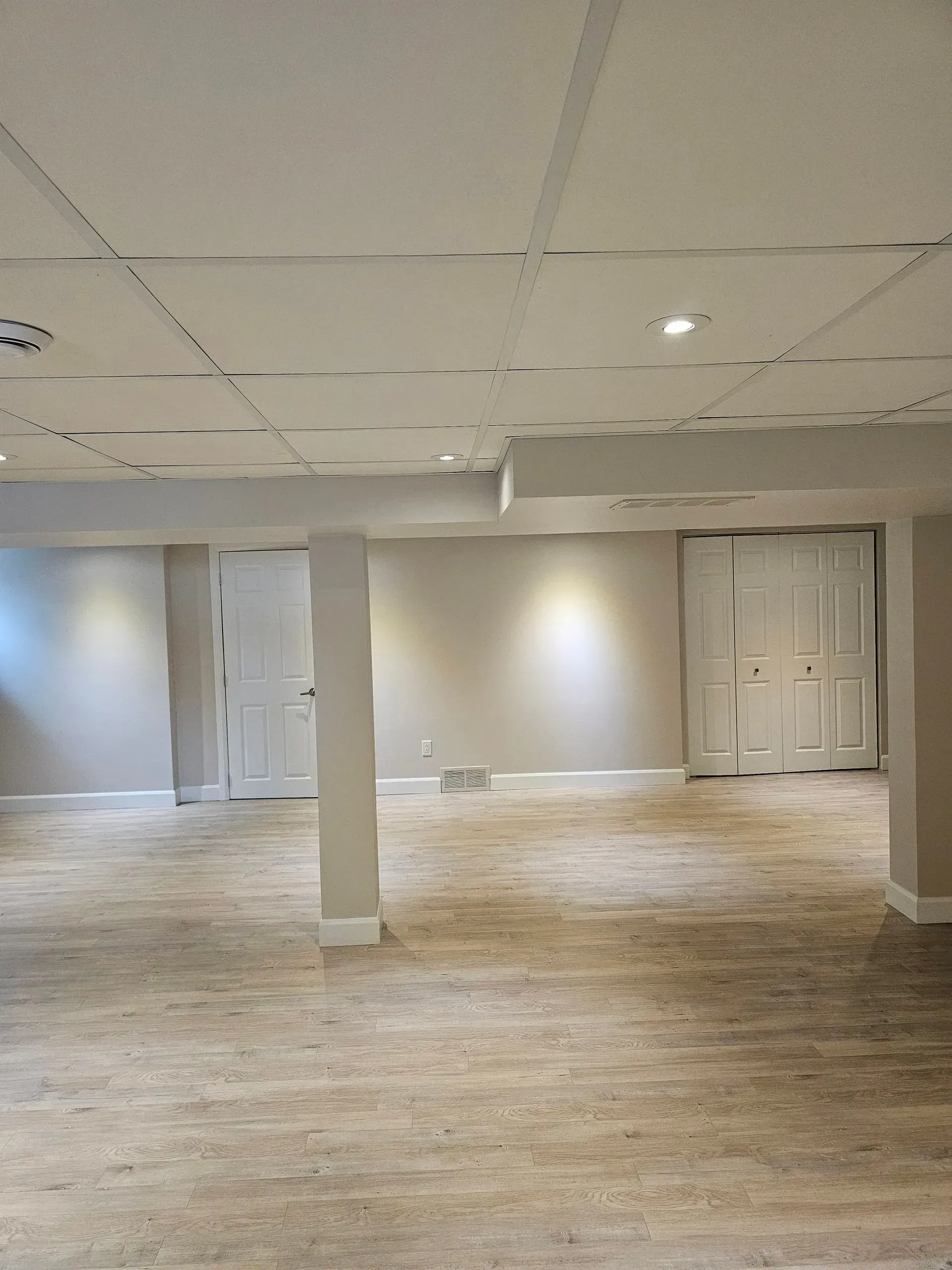 Pièce de sous-sol vide et non aménagée avec des carreaux de plafond blancs, de la moquette beige et deux portes blanches.