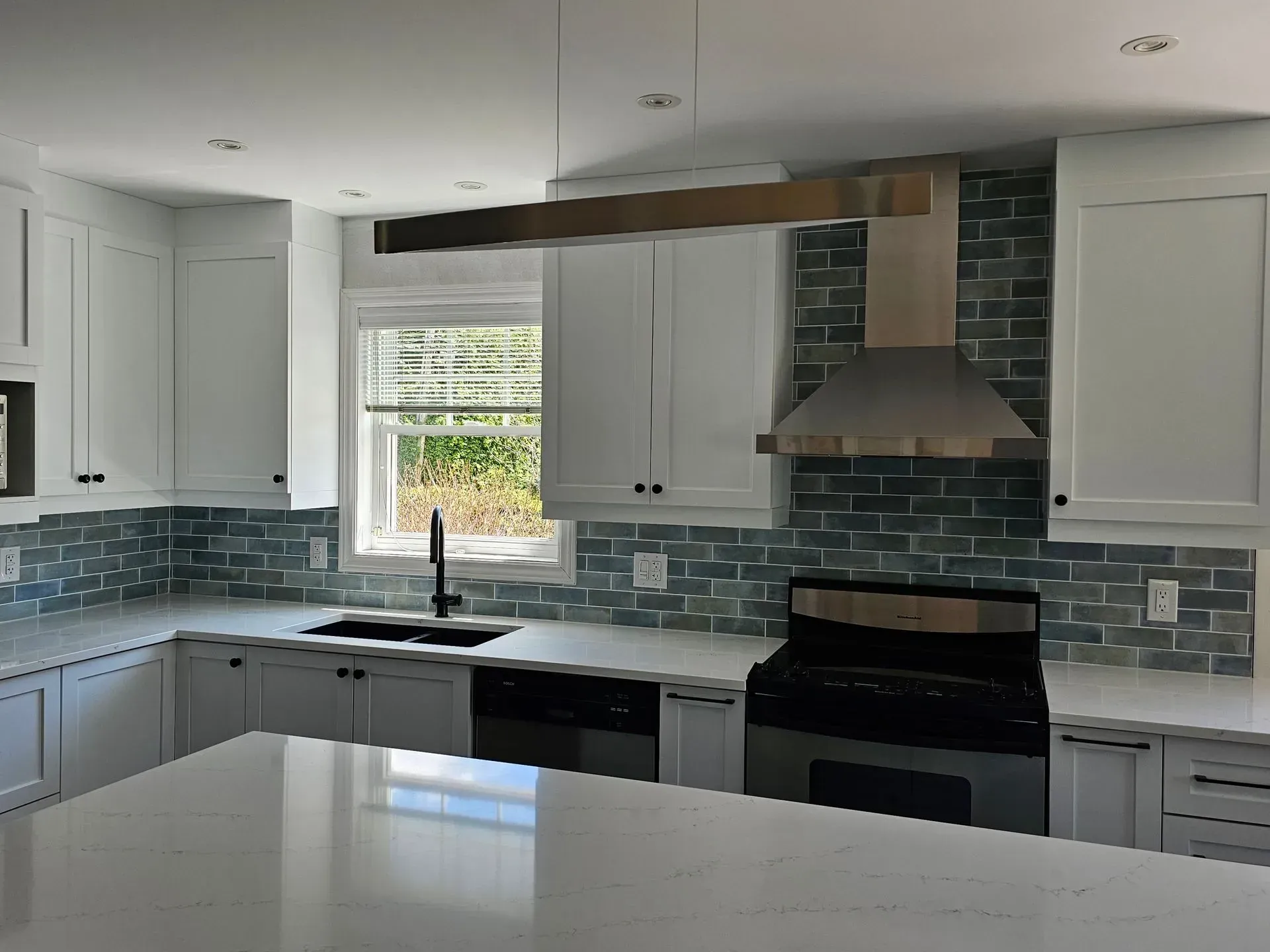 Cuisine moderne et lumineuse avec armoires blanches, crédence en carrelage bleu, grande fenêtre et hotte en acier inoxydable.