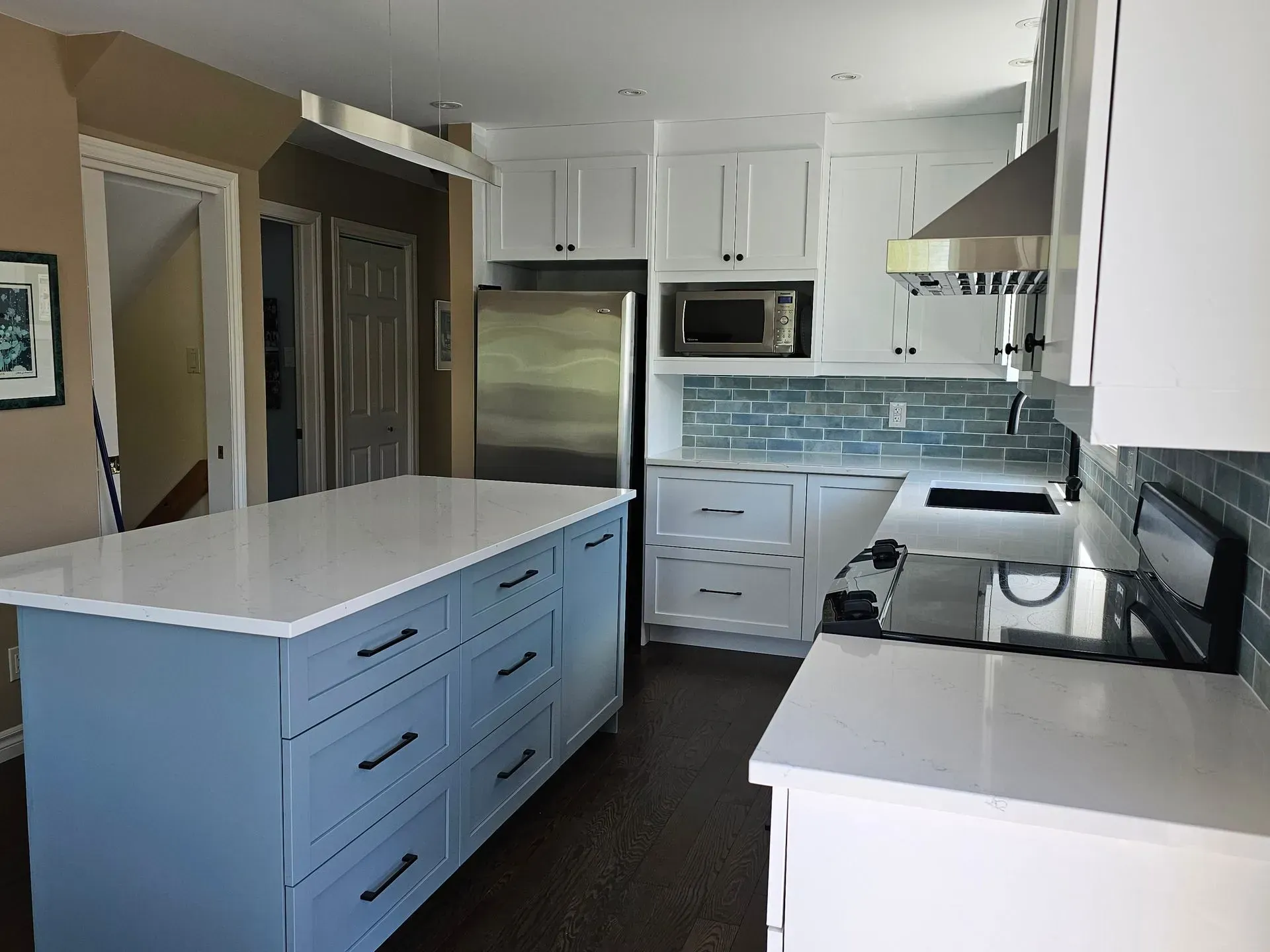 Cuisine blanche lumineuse avec îlot bleu, réfrigérateur en inox et plans de travail noirs.