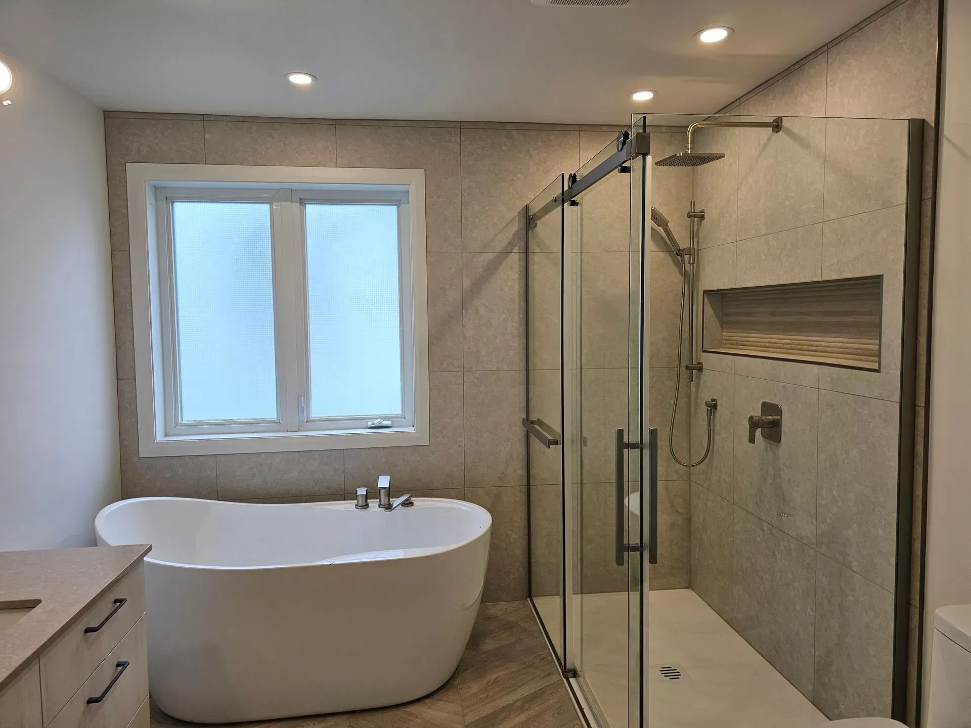 Salle de bains moderne avec baignoire îlot, douche en verre, fenêtre et murs carrelés beiges