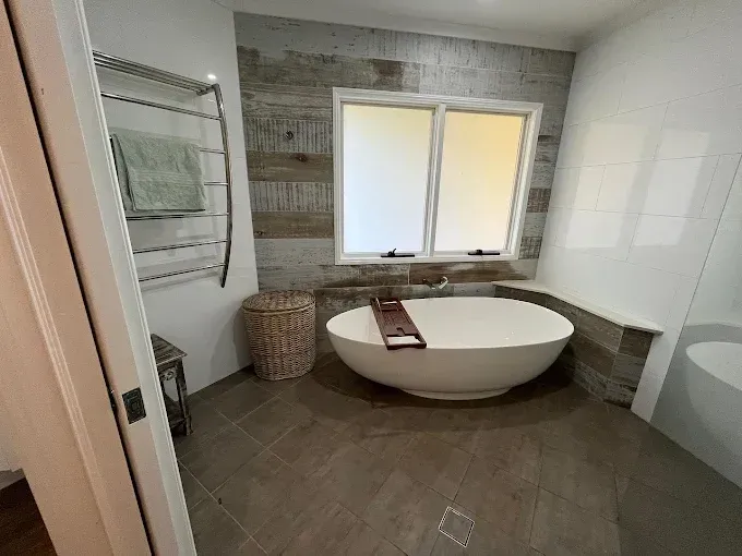 Oval freestanding bathtub against rustic timber wall, with towel warmer and wicker basket. — Rob Powell Plumbing Pty Ltd in Coraki, NSW