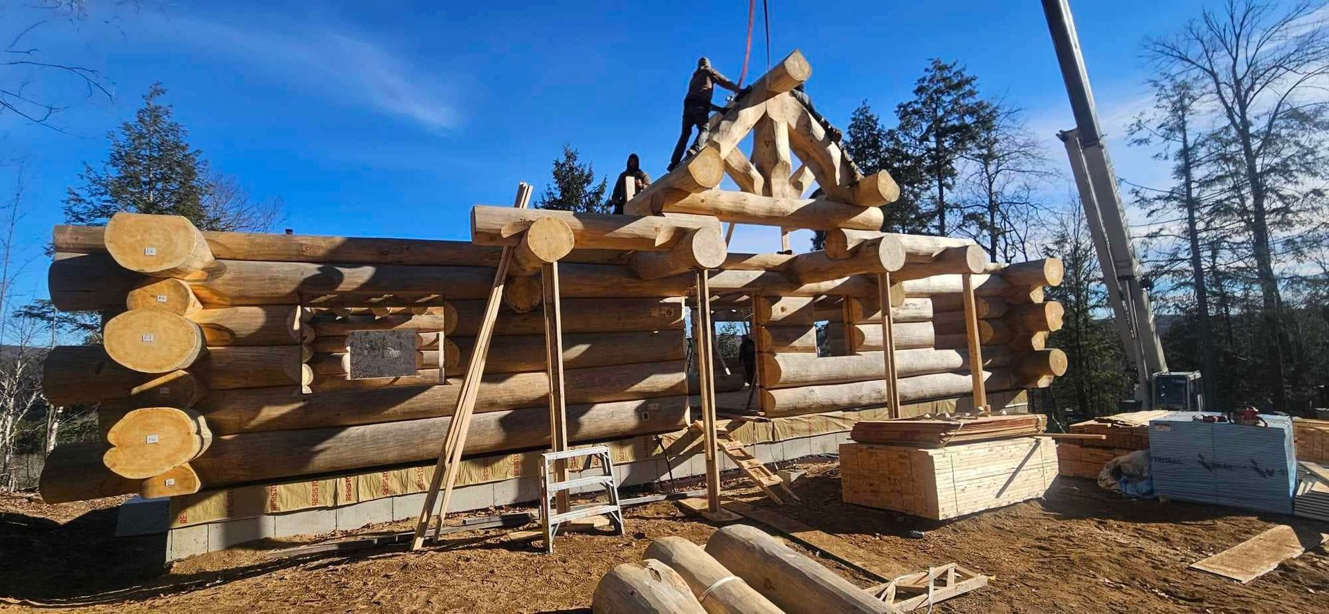 Log cabin under construction with stacked logs, roof trusses, and bright blue sky