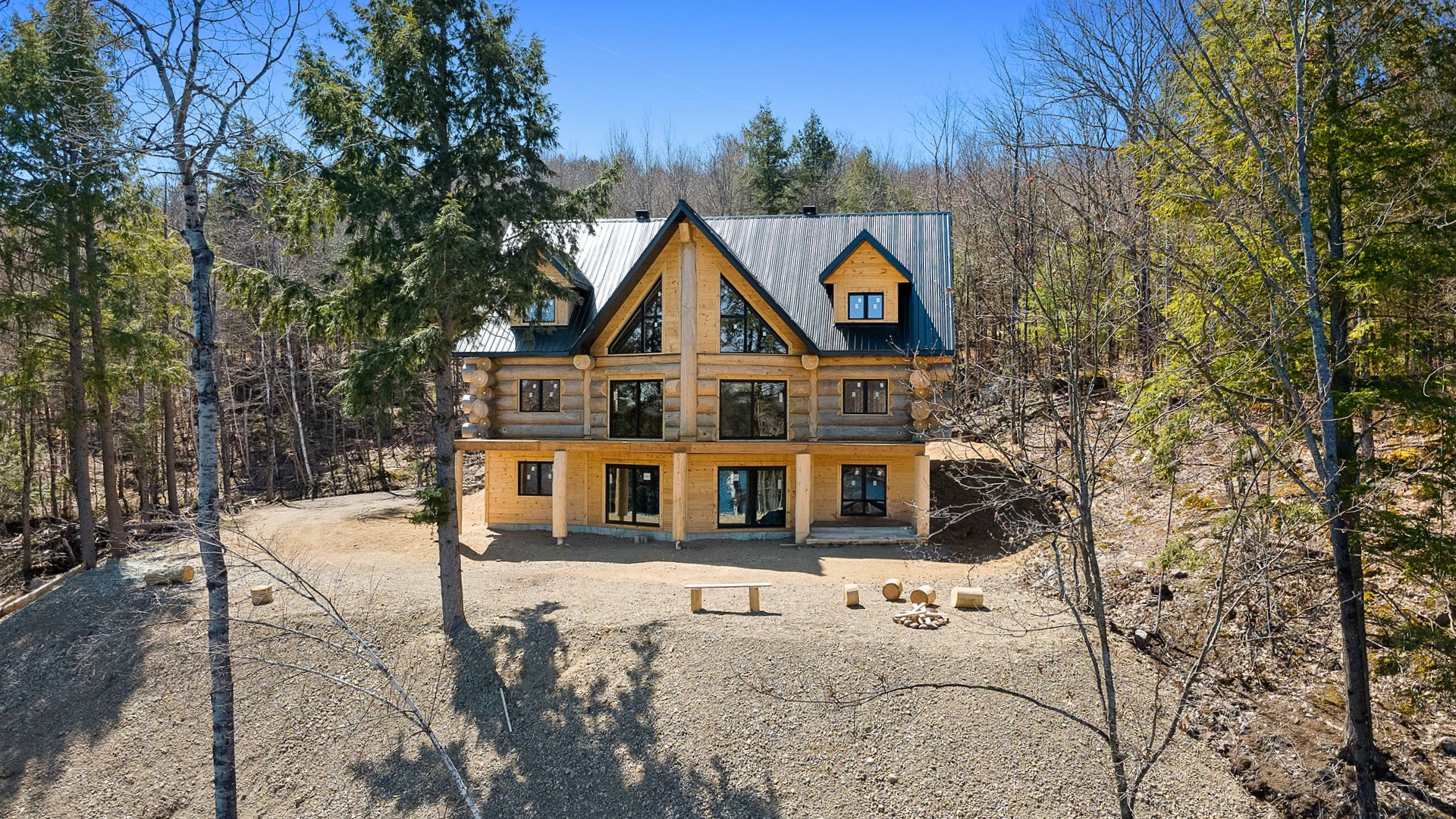 Two-story tan cabin-style house with black roof in a wooded, rocky clearing under a bright blue sky