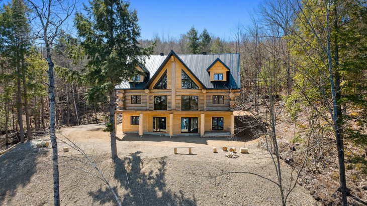 Maison beige au toit noir dans une clairière boisée sous un ciel bleu