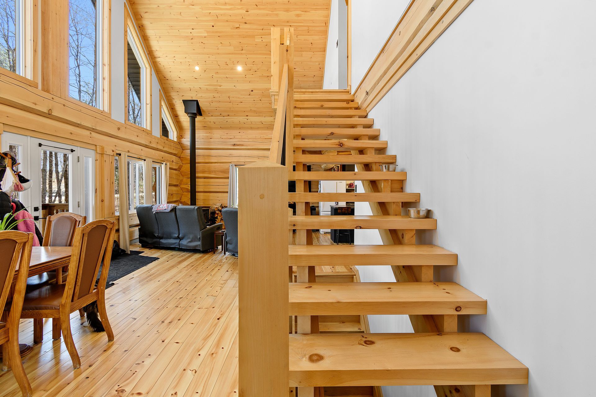 Wooden staircase in a bright log cabin living room with large windows and a dining table nearby