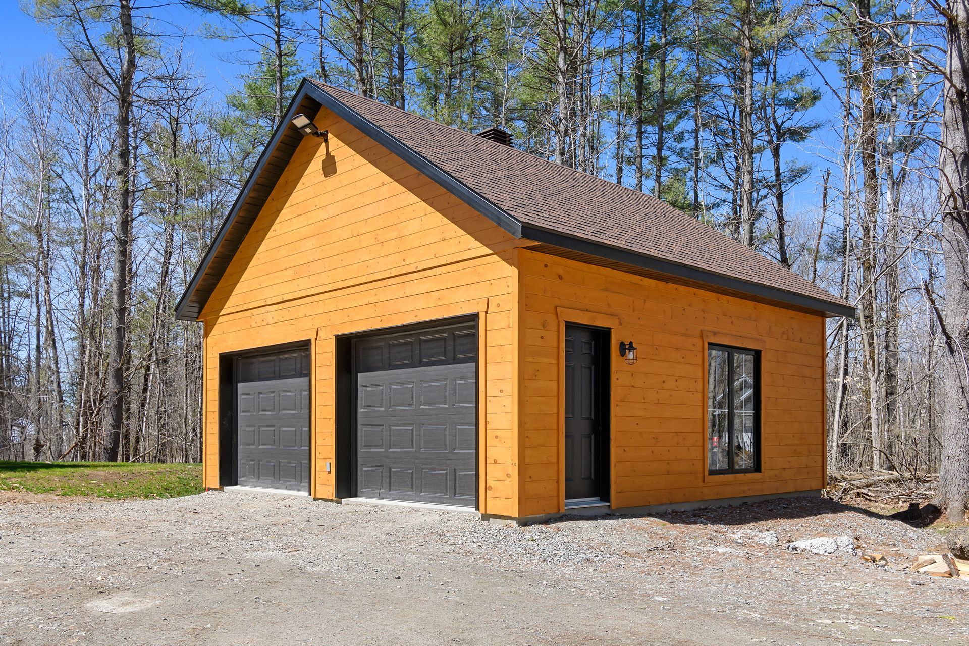 Orange detached garage with two black doors and a side entry, set among trees on a gravel lot.