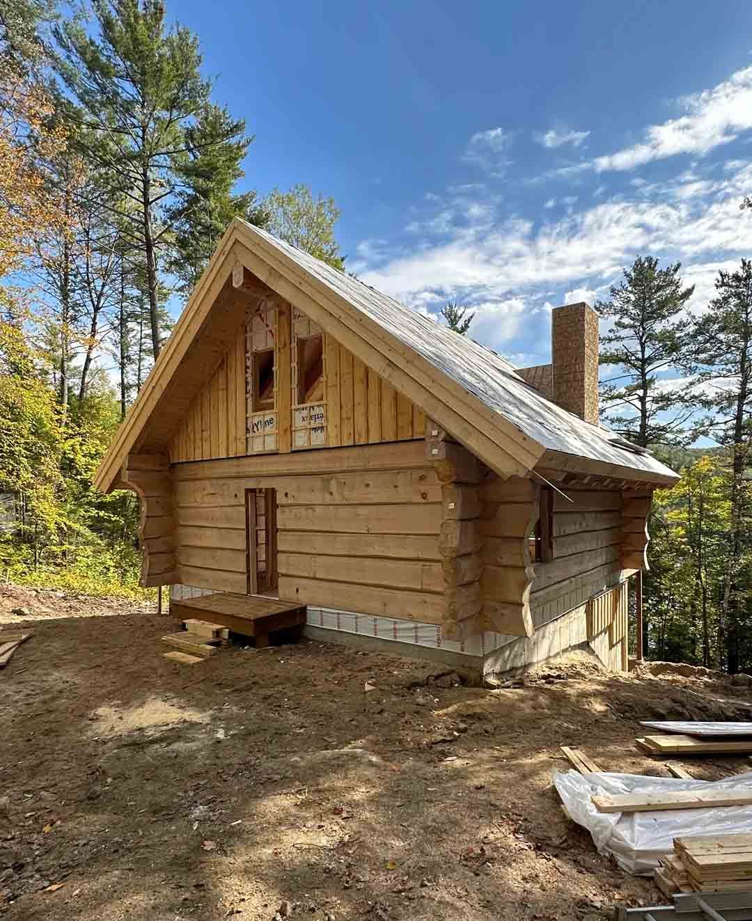 Small unfinished log cabin with steep roof in a wooded clearing under a blue sky