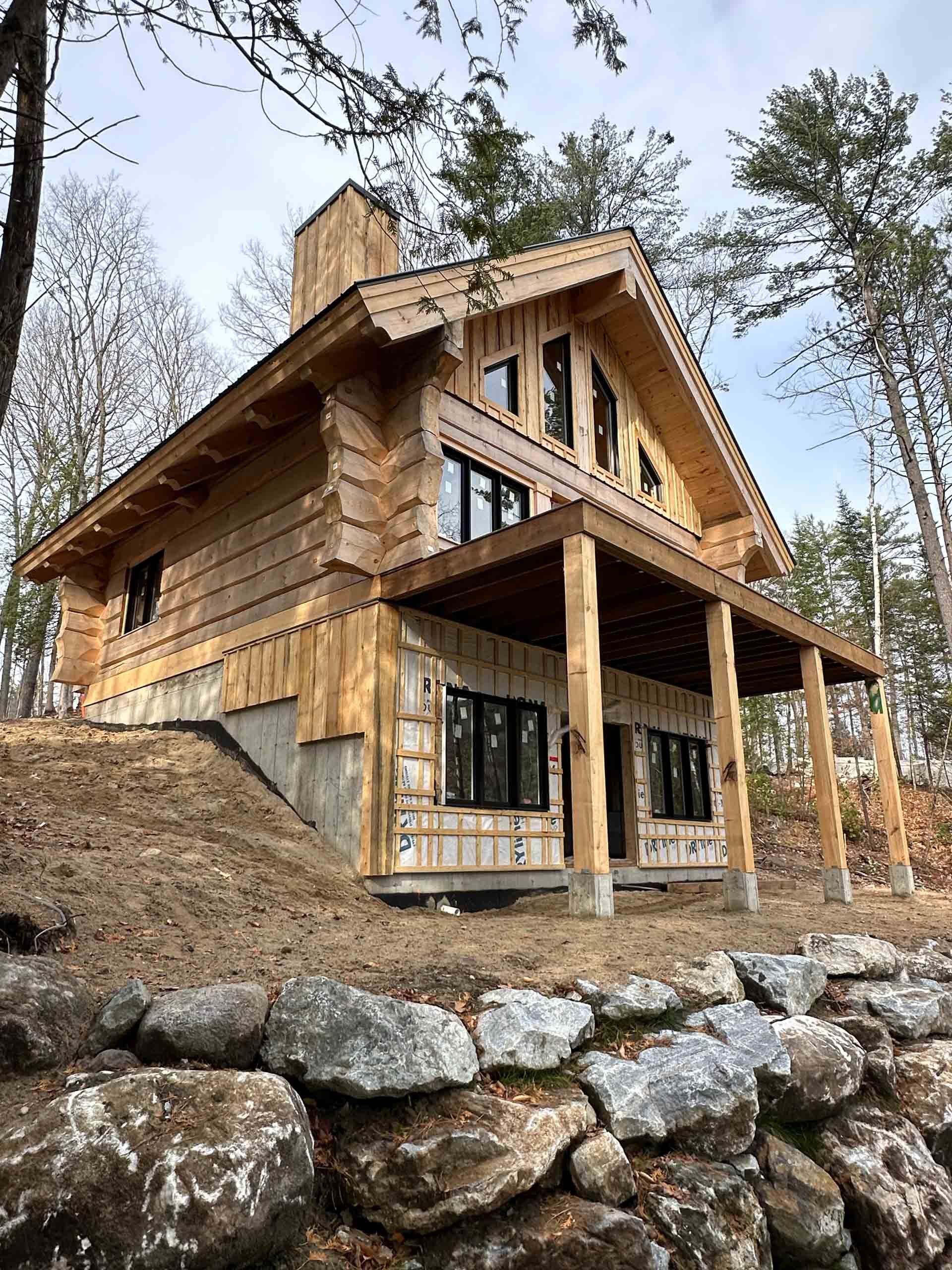 Wooden cabin under construction on a wooded hillside, with stone foundation and large windows.