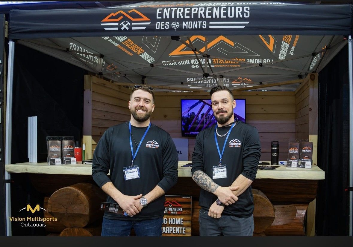 Two men stand behind an entrepreneurs booth with black shirts and a wood-paneled backdrop.