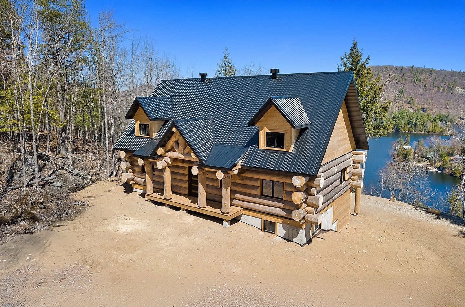 Log cabin with dark roof on a sandy shore beside a lake, surrounded by trees and hills