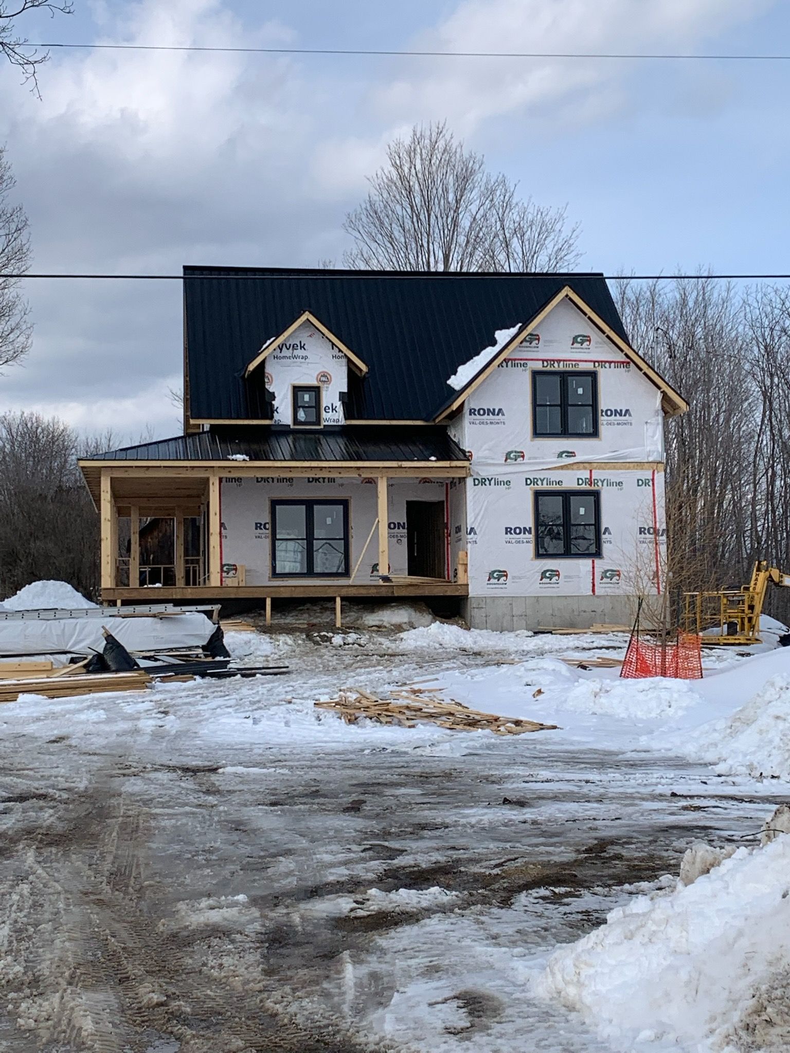 Snow-covered construction site with a partially finished white house and dark roof under a cloudy sky.
