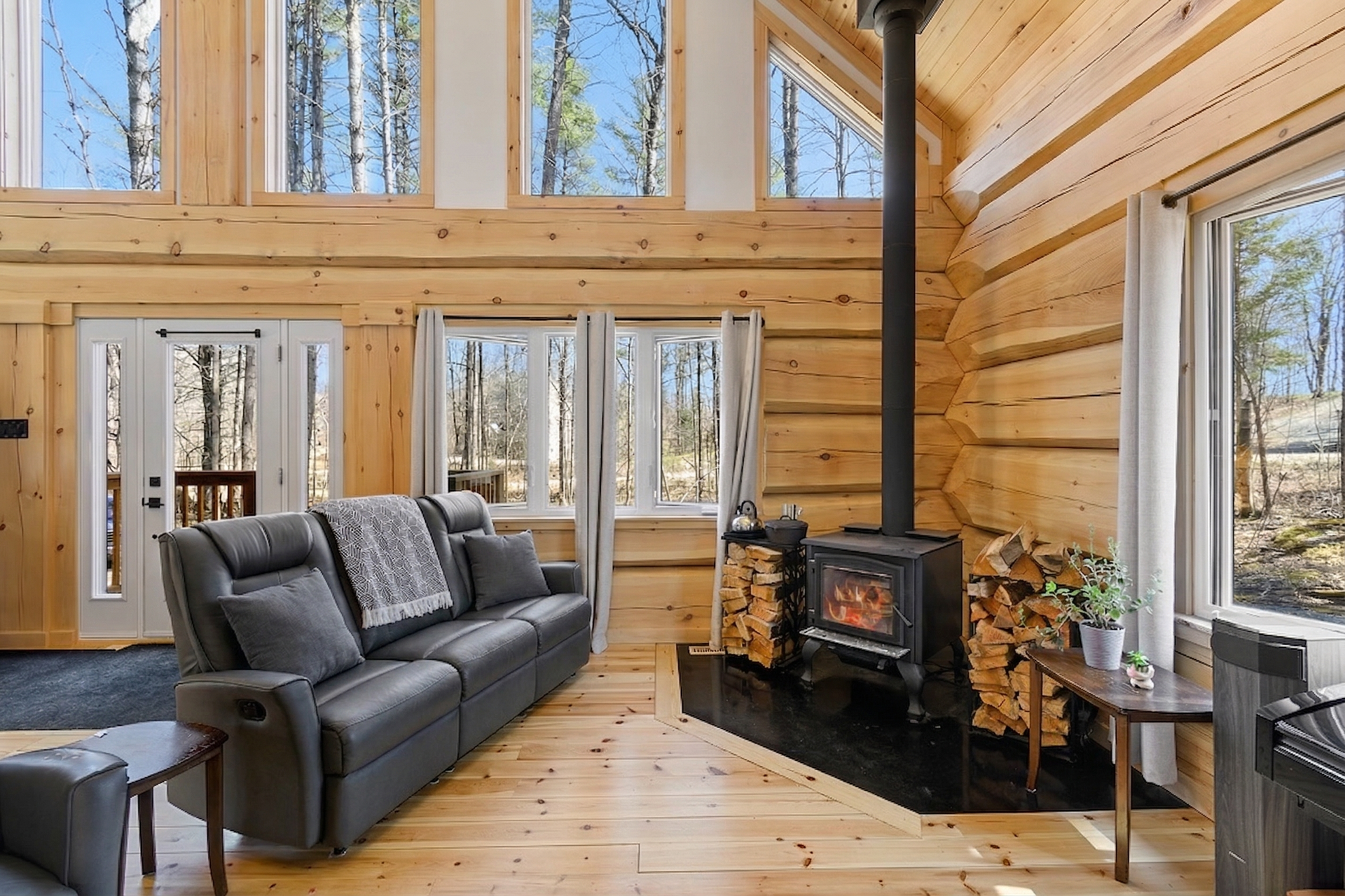 Cozy wood-paneled cabin living room with gray sofa, large windows, and a black wood stove.