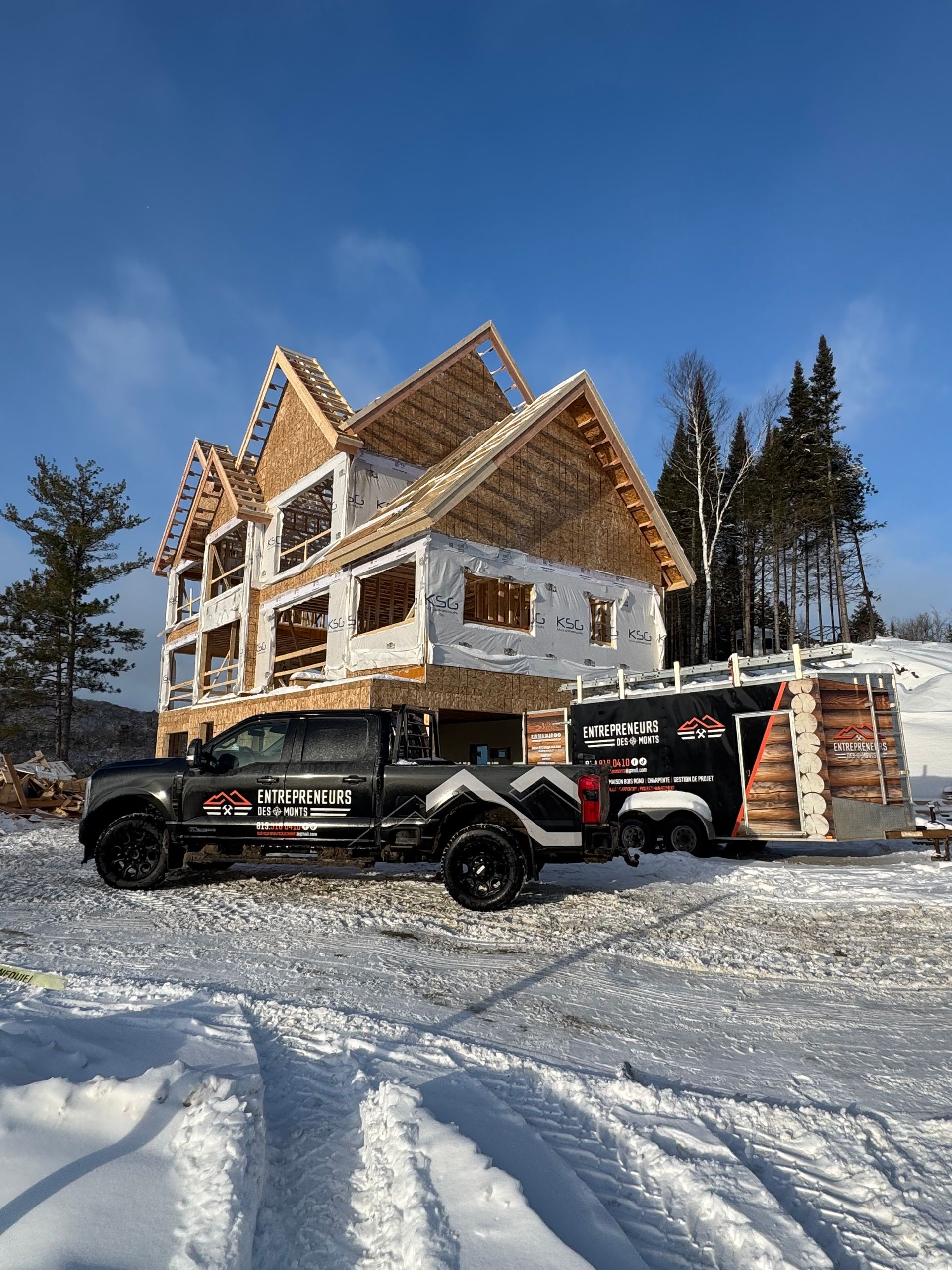 Snowy construction site with a large wooden house under construction and two black utility vehicles in front
