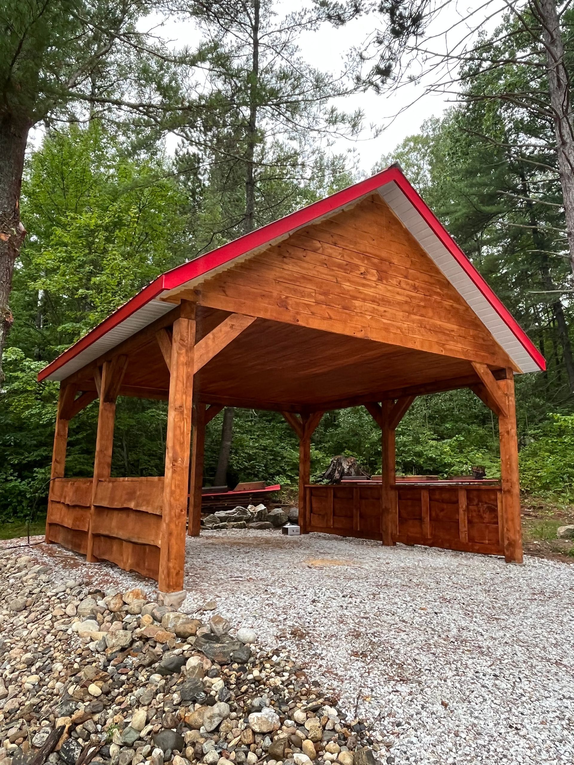 Wooden pavilion with red roof and stone base in a wooded outdoor setting