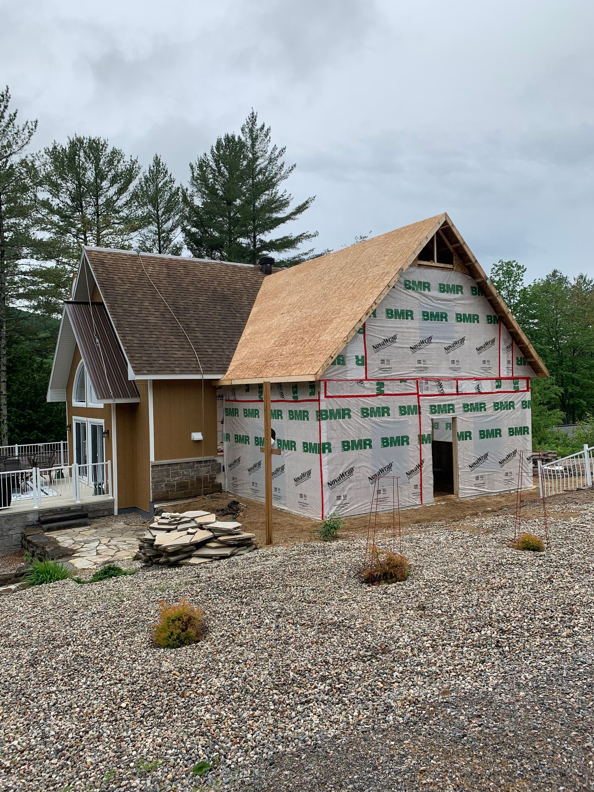 Partially built house with exposed wood framing, wrap, and shingle roof on a gravel lot under cloudy sky