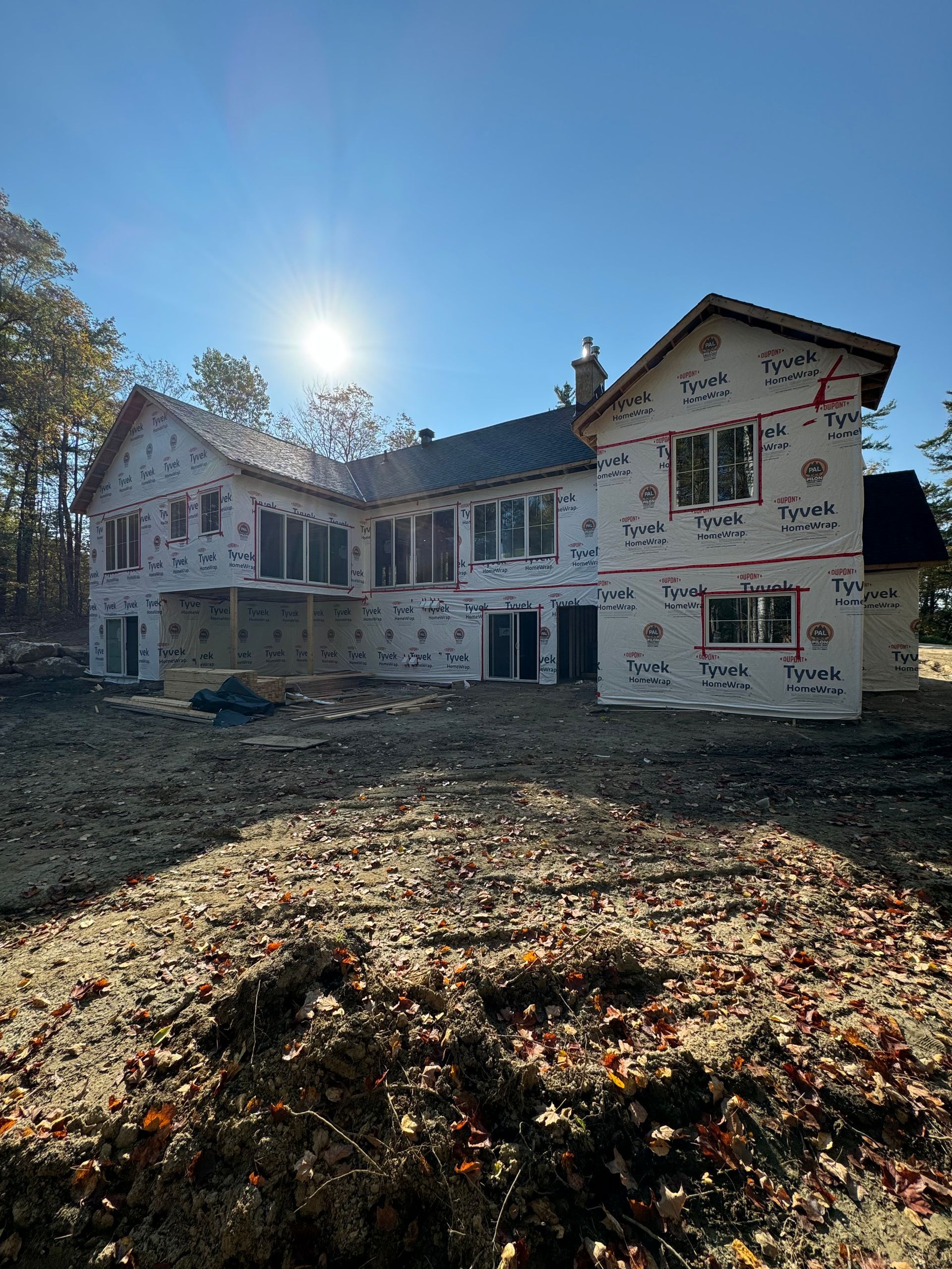 Partially built house with white siding and exposed framing under a bright sun, surrounded by leaves