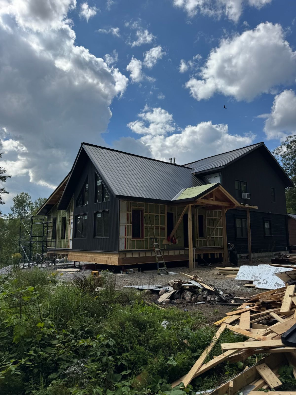 Partially built dark house with metal roof and debris in front under a cloudy blue sky